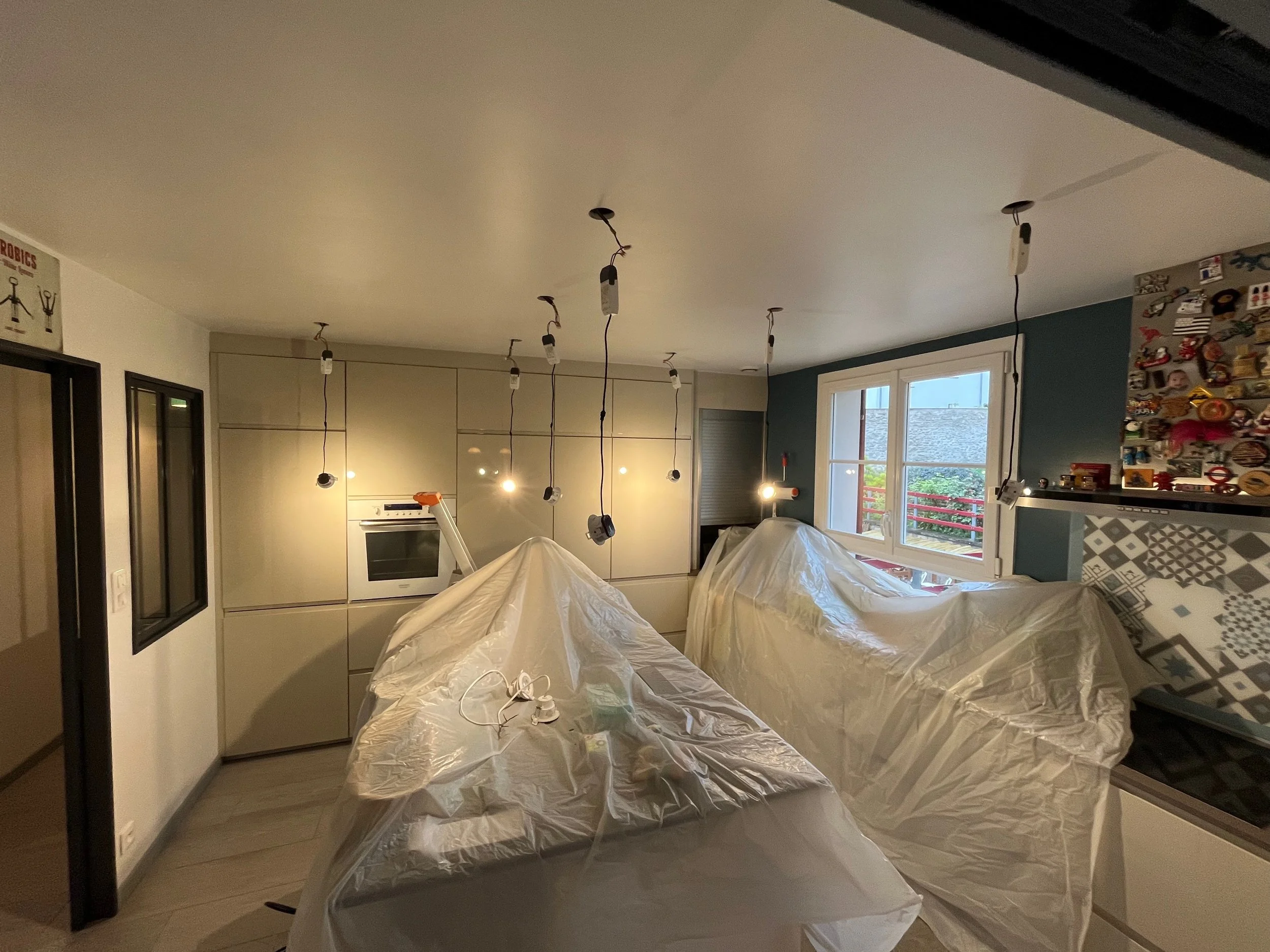 Une cuisine moderne en cours de rénovation avec des lumières suspendues non installées et des meubles recouverts de plastique.