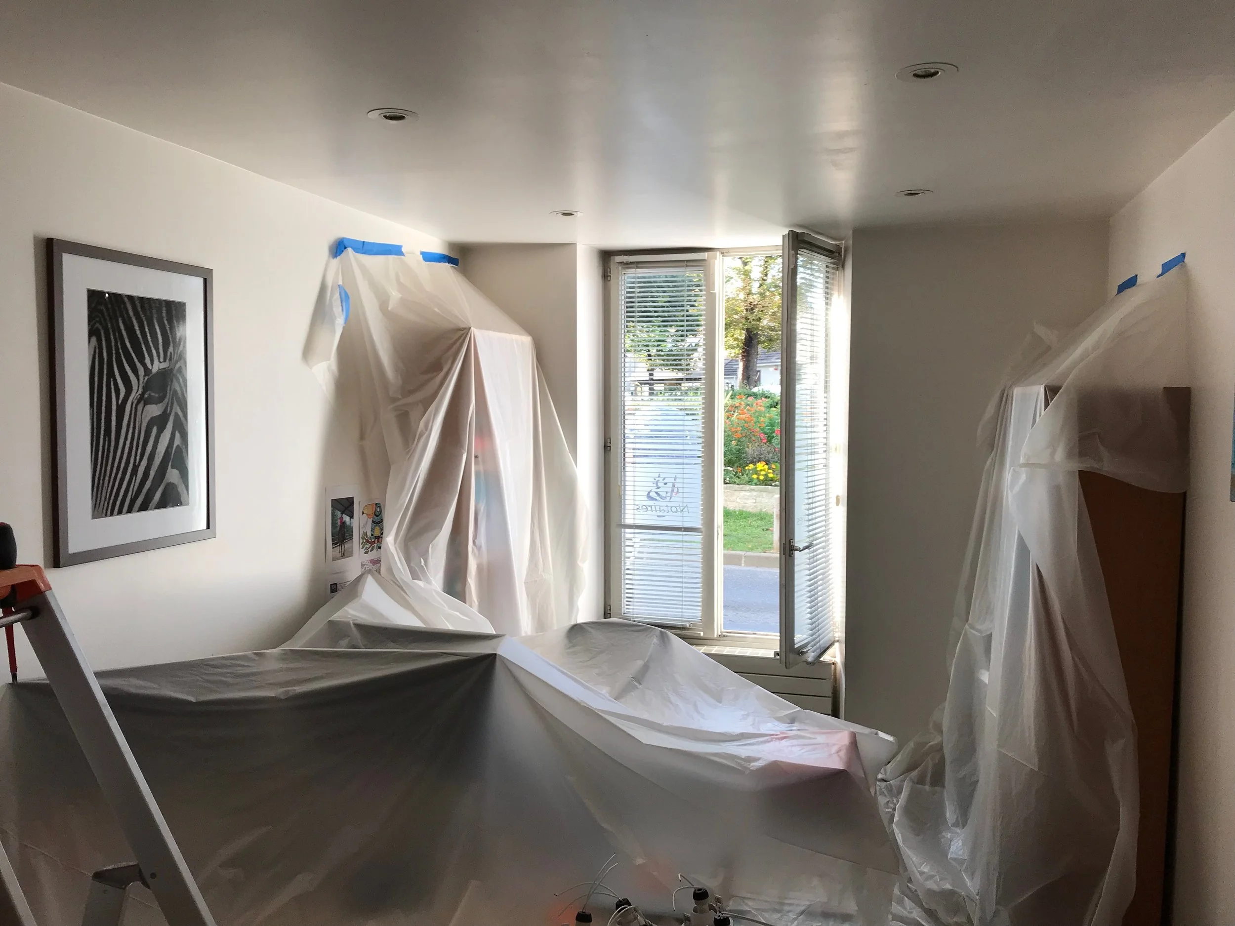 Intérieur d'une pièce en rénovation, protégée par des draps en plastique, avec un lit et un bureau, fenêtre ouverte laissant entrer de la lumière naturelle.