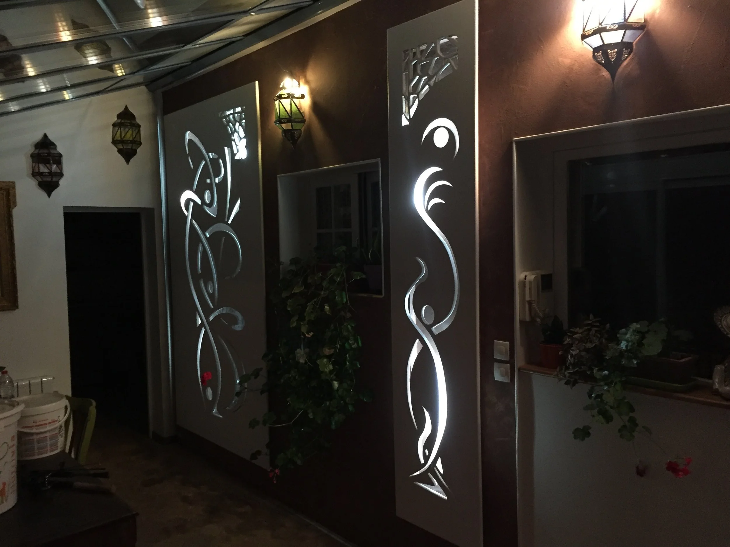 Intérieur d'une pièce avec deux portes éclairées avec des motifs lumineux stylisés, lampes murales en forme de lanterne, plantes et une fenêtre.