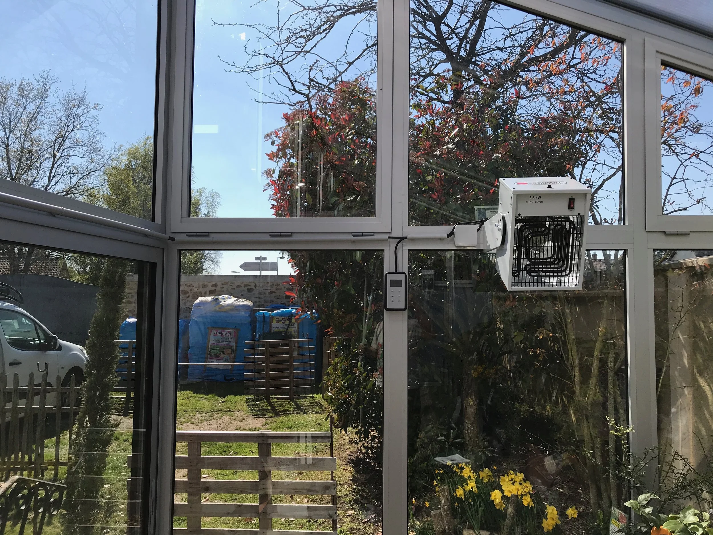 Véranda avec un ventilateur électrique blanc fixé à la fenêtre et un panneau de contrôle, vue sur un jardin ensoleillé avec des plantes, un arbre avec des feuilles rouges et des objets stockés dehors.