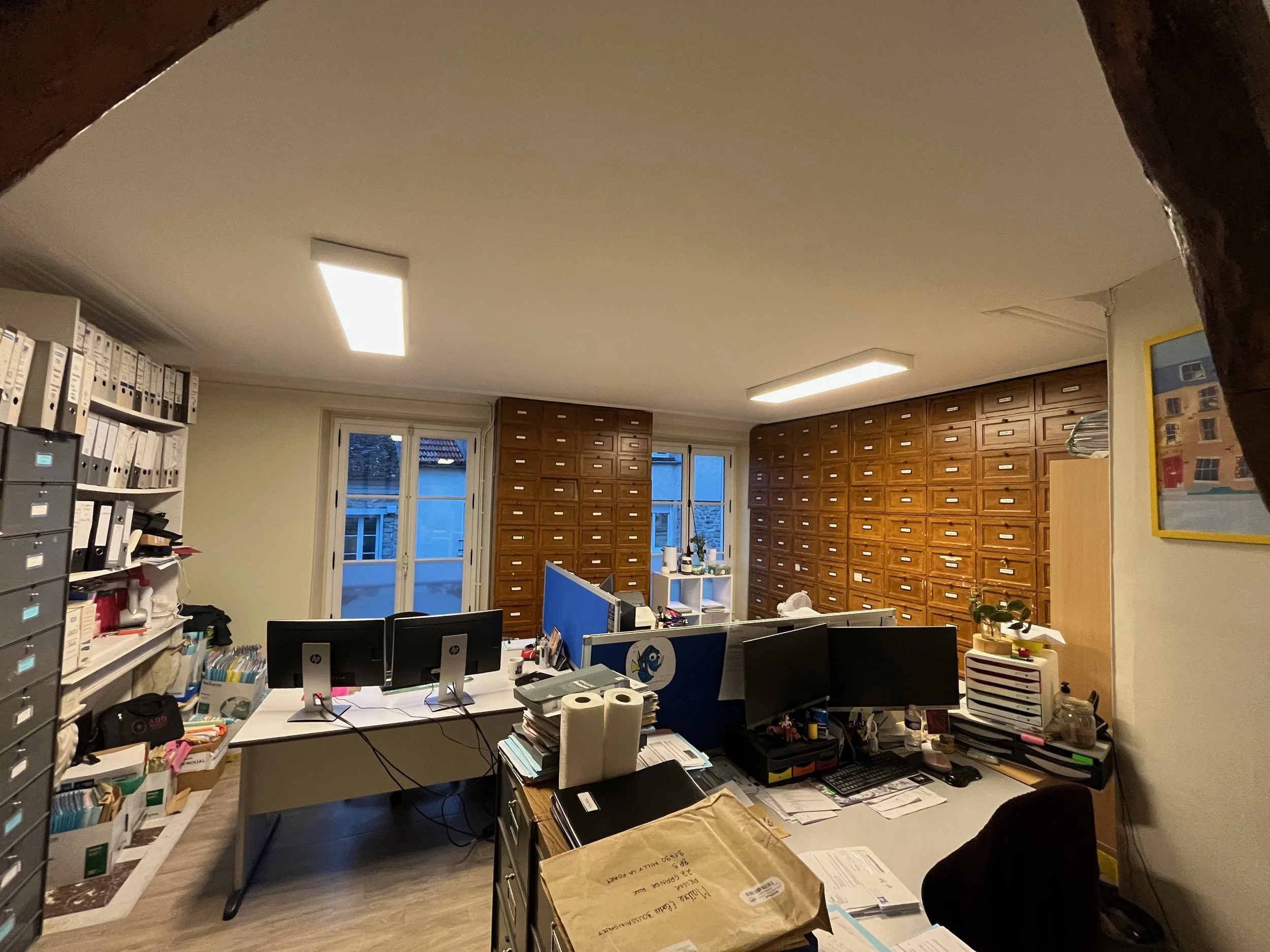 Bureau avec plusieurs ordinateurs, étagères pleines de classeurs, et mobilier de bureau, éclairé par des lumières de plafond, avec deux fenêtres en arrière-plan.