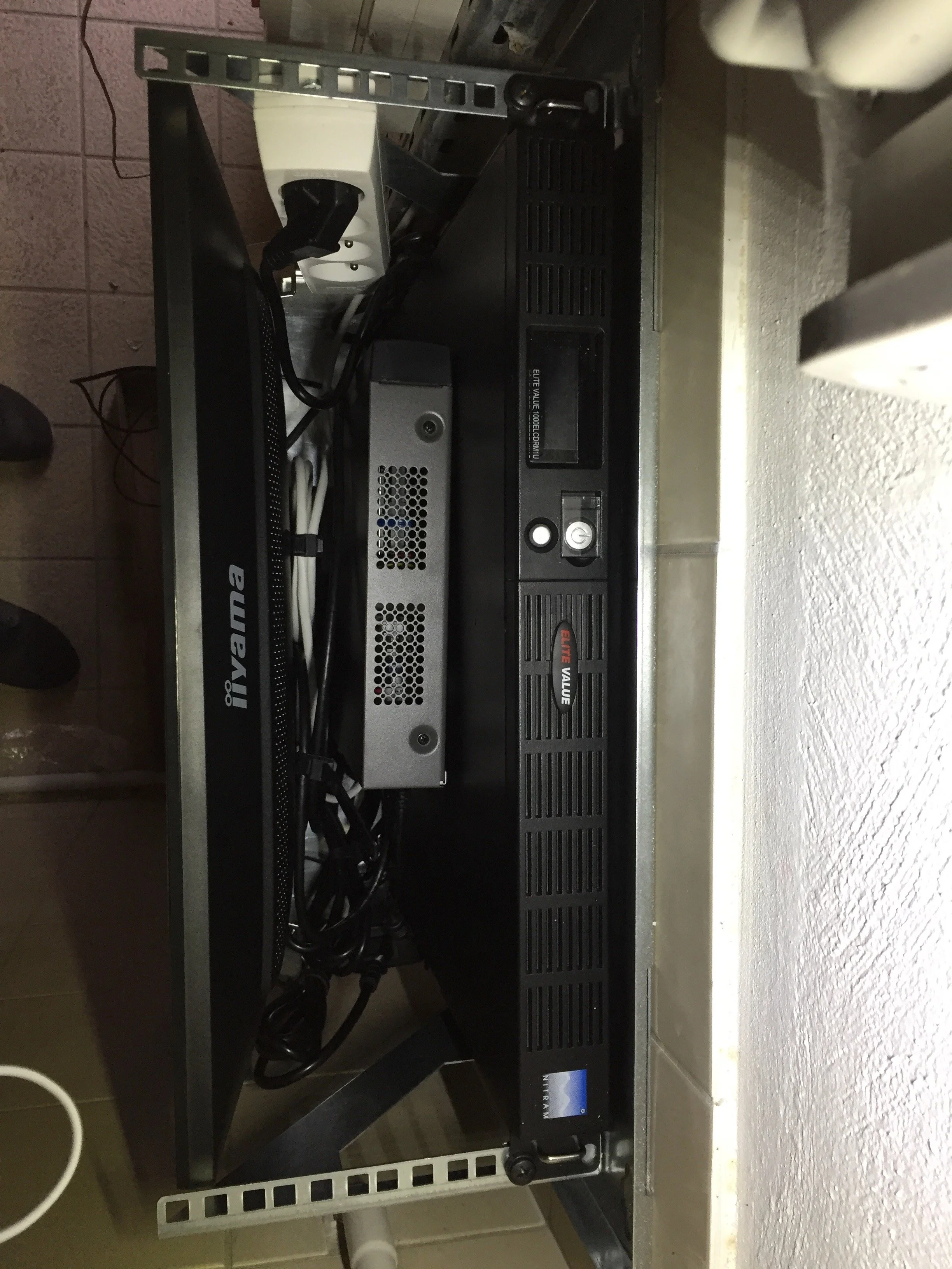 Équipement électronique sur une étagère métallique, incluant un ordinateur ou un appareil de serveur, un câble d'alimentation, et autres composants électroniques.