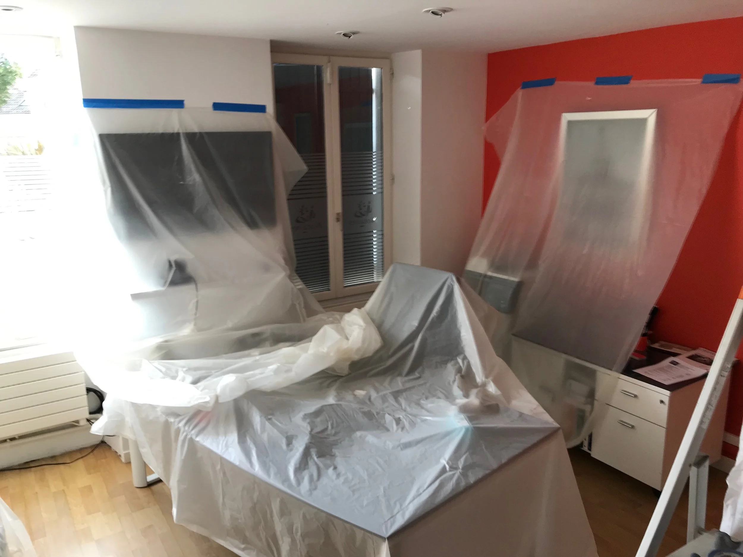 Une pièce en cours de peinture avec du plastique de protection sur les murs, un radiateur et une table recouverte d'une bâche blanche, fenêtres en arrière-plan.