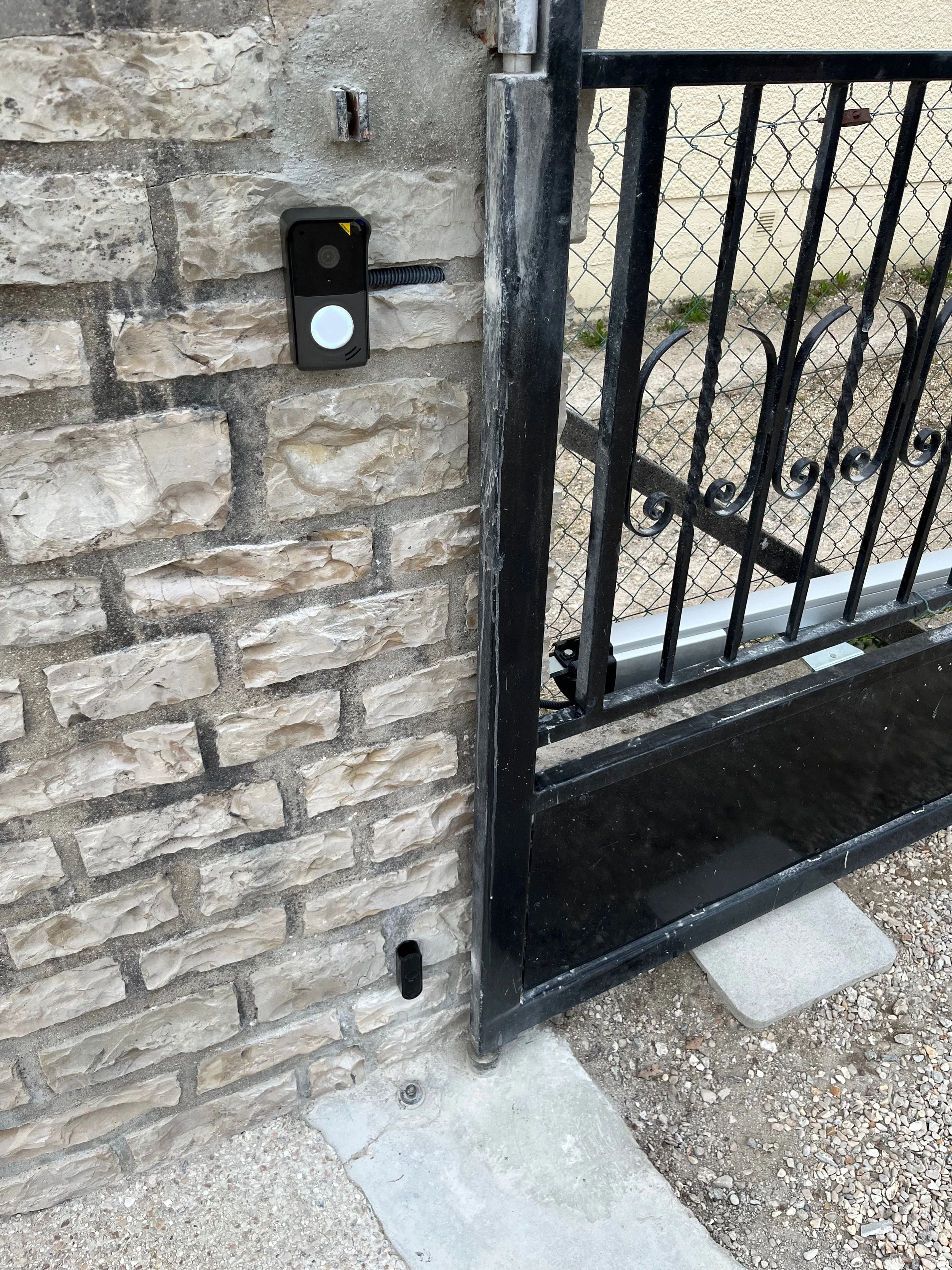 Véranda avec un portail en métal noir équipé d'un système d'ouverture automatique ou électrique, côté extérieure d'un mur en pierre avec un interphone ou un lecteur de badge.