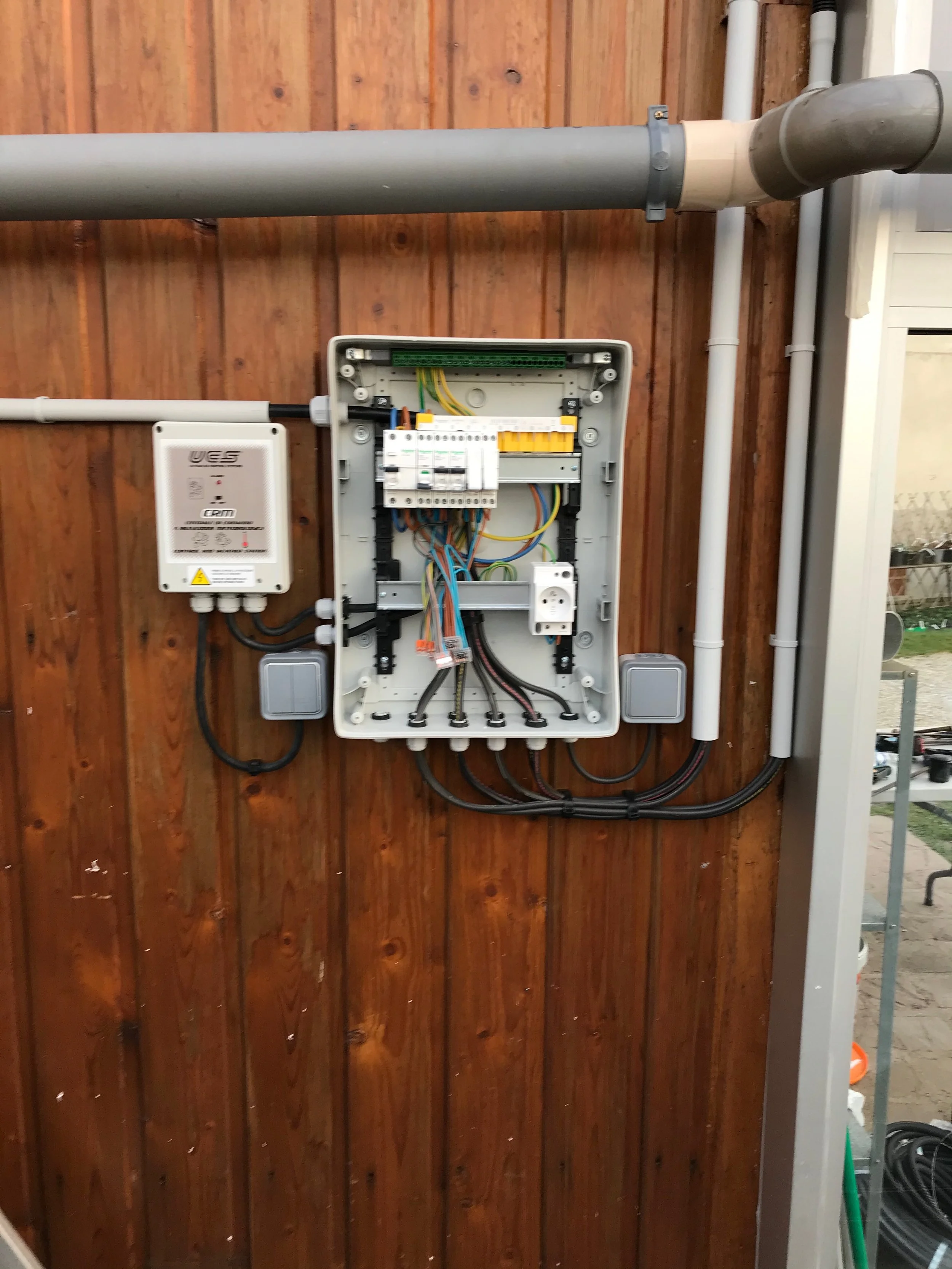 Un boîtier électrique ouvert avec des fils colorés, des disjoncteurs et des composants électroniques, fixé à un mur en bois.