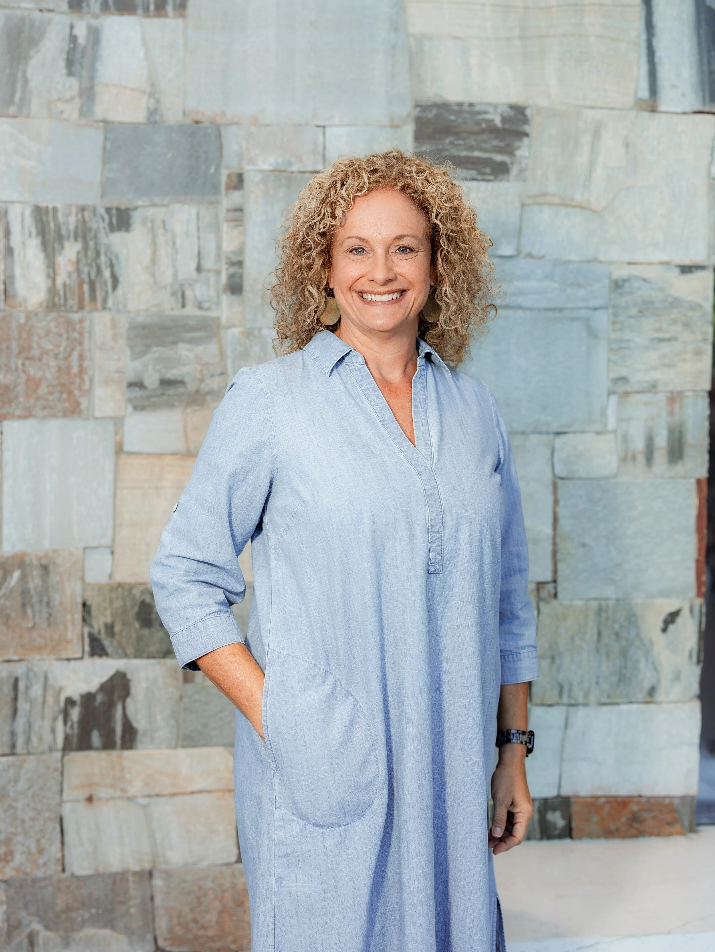 A woman with curly blonde hair smiling, wearing a pink knitted sweater, in a dimly lit room with blurred lights and objects in the background.