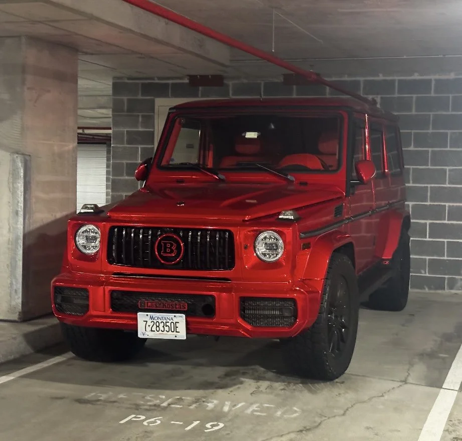 A red luxury SUV, specifically a Brabus G-Class, parked in an underground garage near a concrete wall and support pillar.