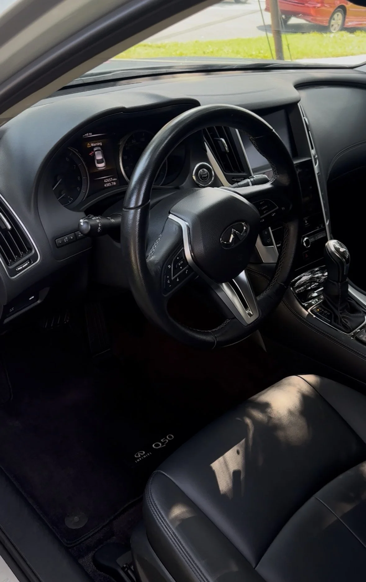 Interior of a luxury car showing the dashboard, steering wheel with Infiniti logo, gear shift, and an Infiniti Q50 branded floor mat.