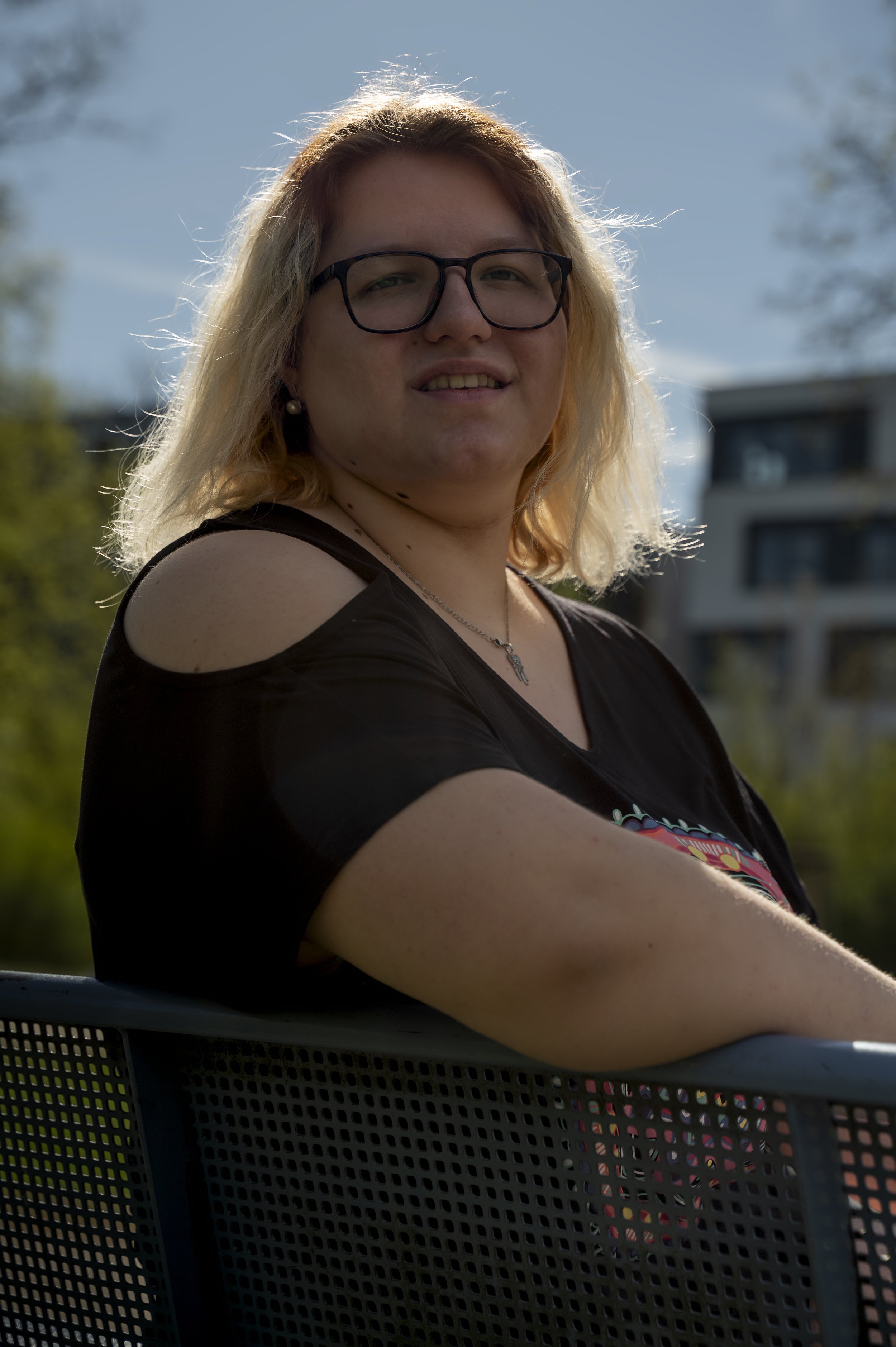 Junge Frau mit Brille, blonde Haare, in schwarzes Top mit Schulterausschnitt, sitzt auf einer Bank im Freien, im Hintergrund Gebäude und Bäume, sonniger Tag.