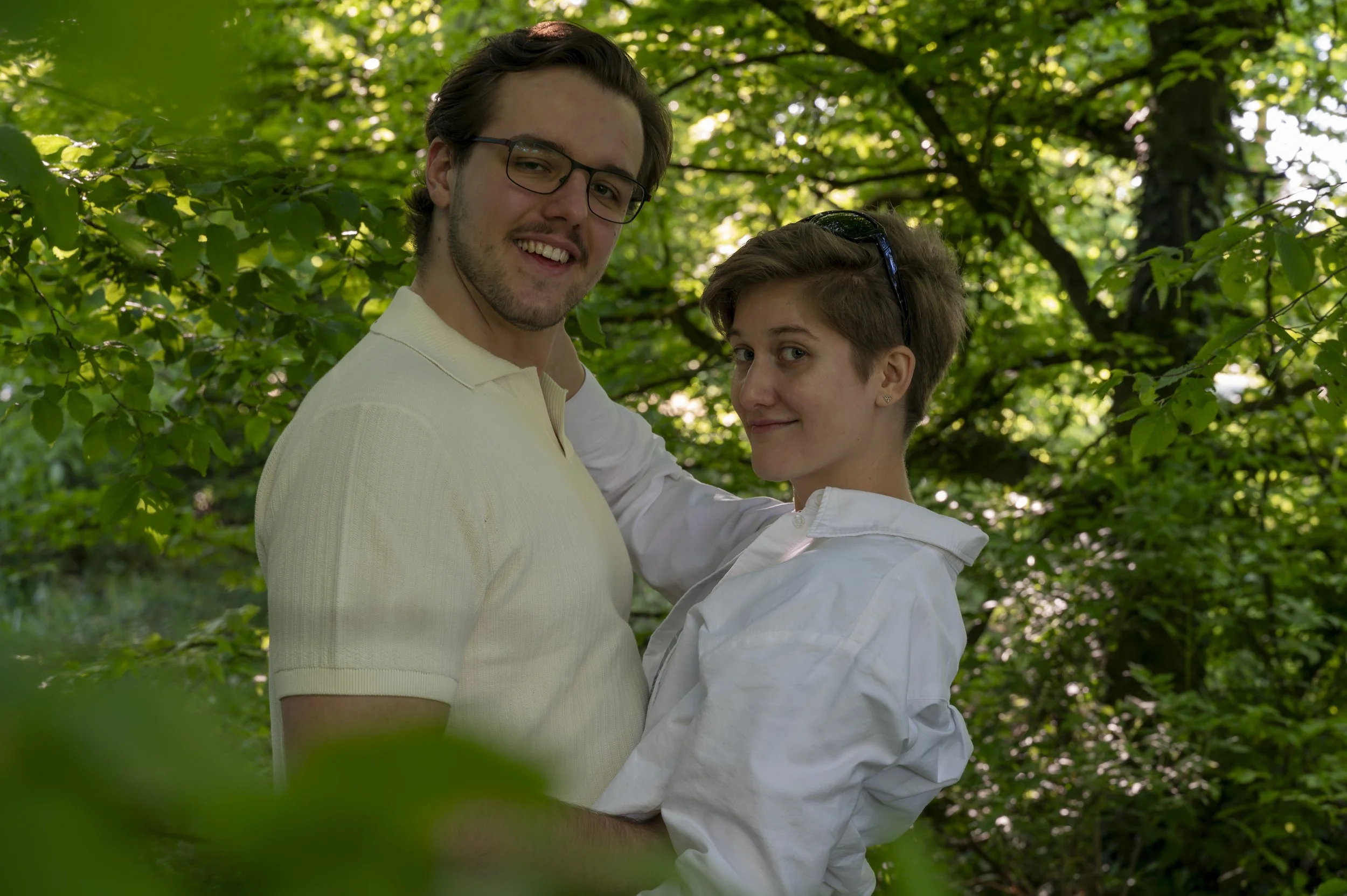 Ein junger Mann und eine junge Frau umarmen sich in einem grünen, bewaldeten Bereich. Sie lachen und schauen in die Kamera, die Szene ist sonnig.