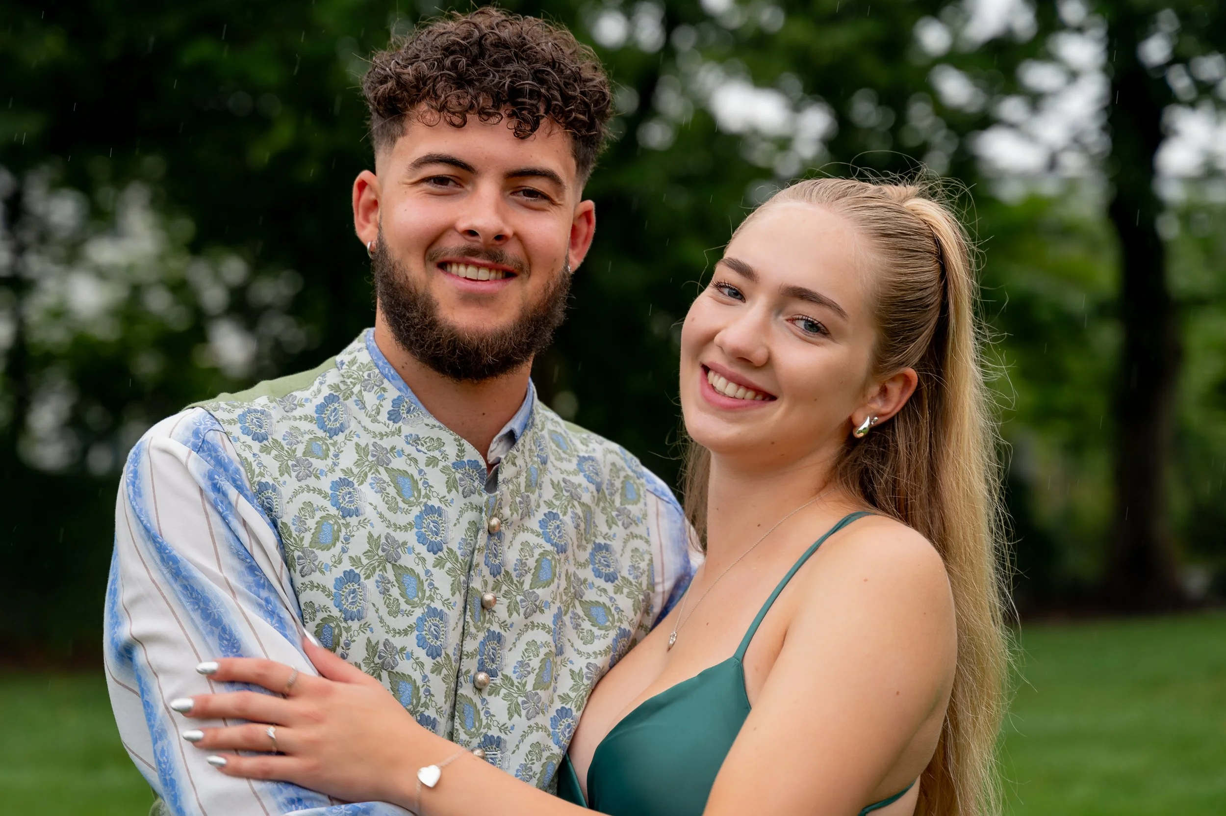 Ein verliebtes Paar lächelnd im Park, die Frau trägt ein grünes Kleid und hat langes, blondes Haar; der Mann trägt ein Blumenmuster-Shirt und hat braune, lockige Haare und einen Bart.