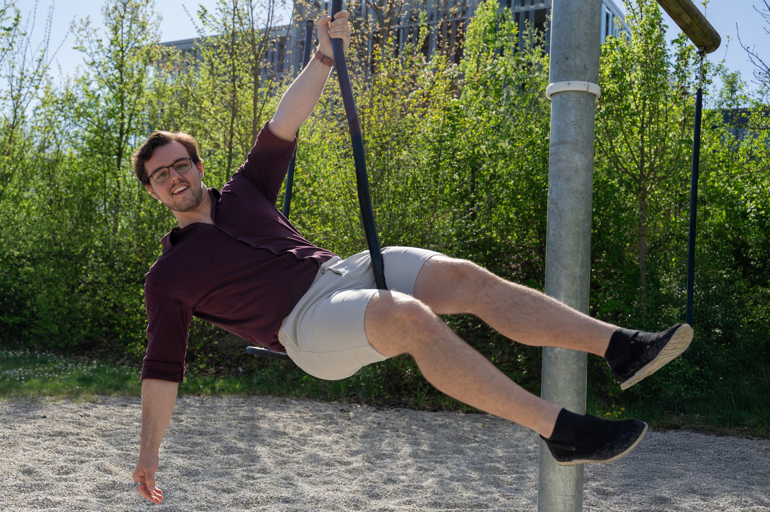 Junger Mann hängt an einer Schaukel im Freien, umgeben von grünen Büschen und Bäumen, bei sonnigem Wetter.