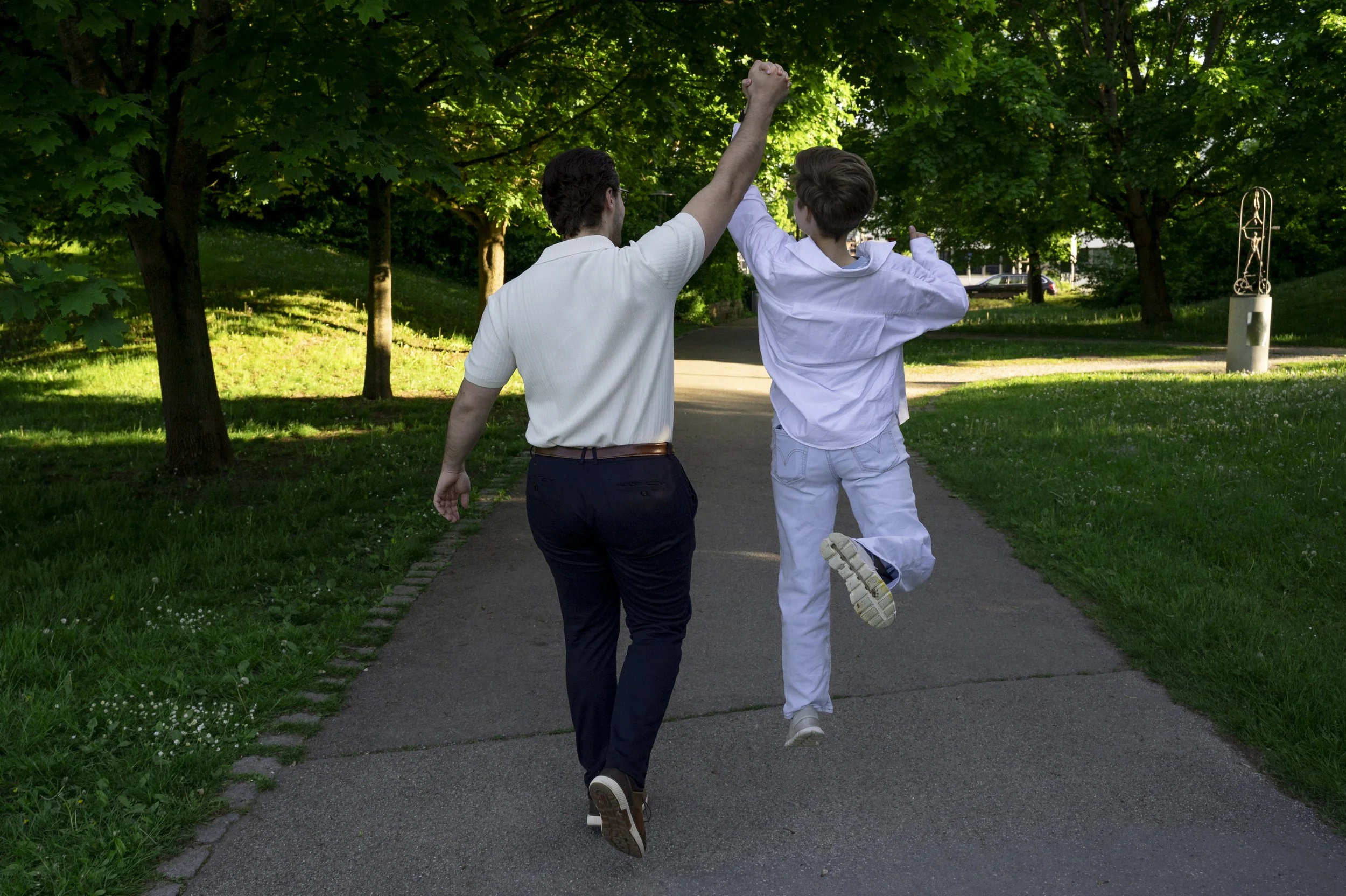 Zwei Männer laufen arm in arm durch einen Park im Sonnenschein, einer hebt den Arm in die Luft.