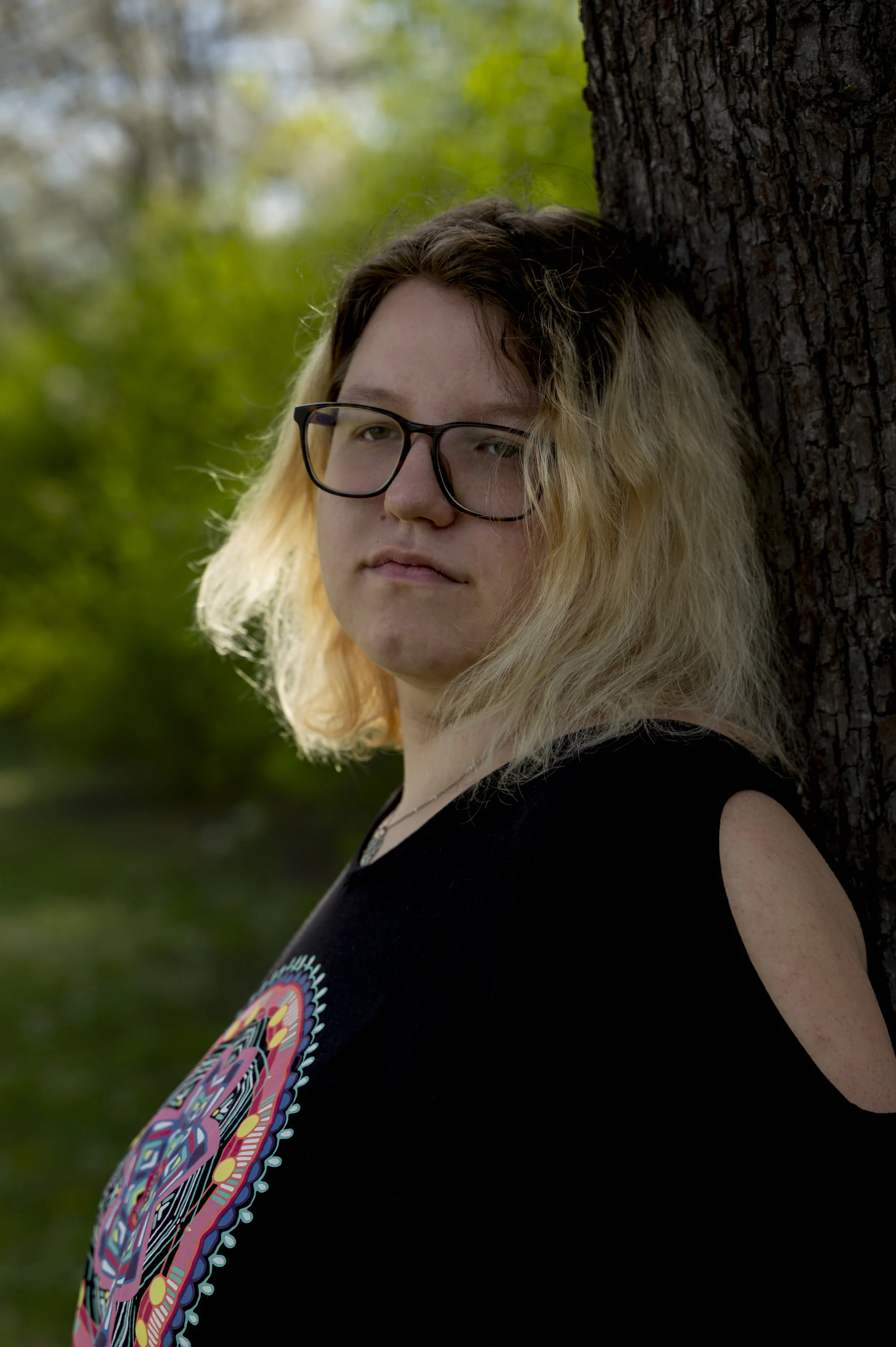 Junge Frau mit blonden, gewellten Haaren, trägt eine schwarze Top mit buntem Mandala-Design und trägt eine schwarze Brille, lehnt an einen Baum im Grünen.