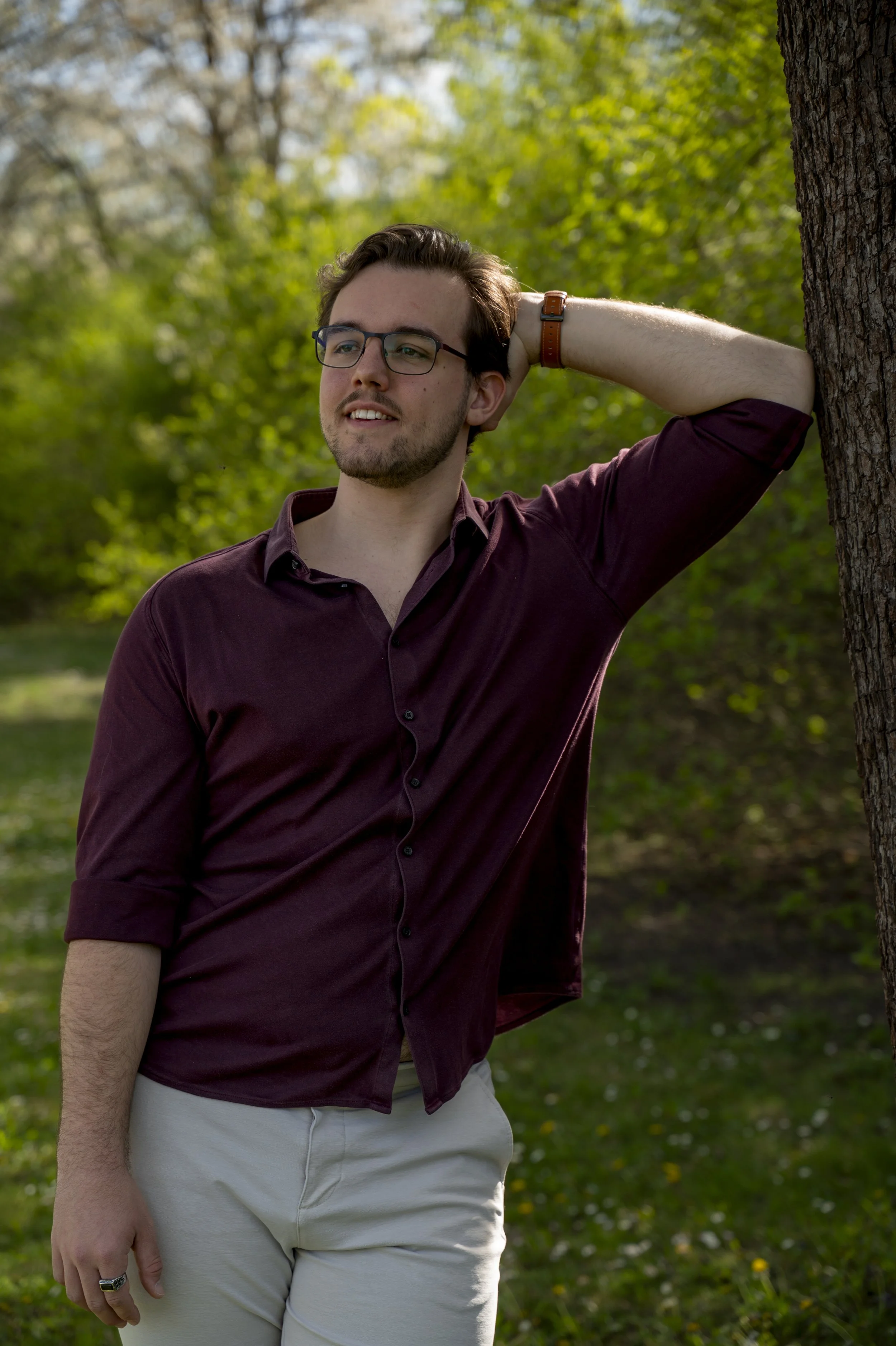 Ein junger Mann mit Brille lehnt an einem Baum im Grünen, trägt ein dunkelrotes Hemd und helle Hose, bei schönem Wetter.