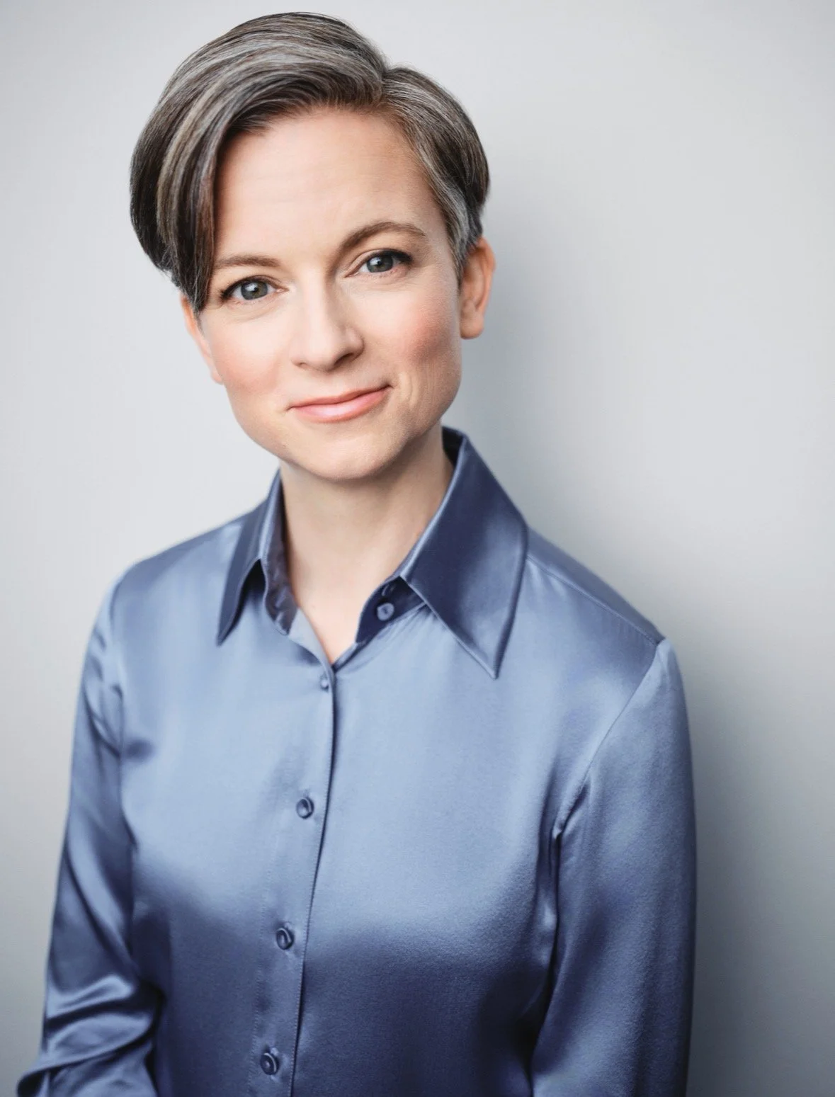 Portrait of a woman with short gray hair, wearing a blue satin shirt, smiling gently, against a plain light gray background.