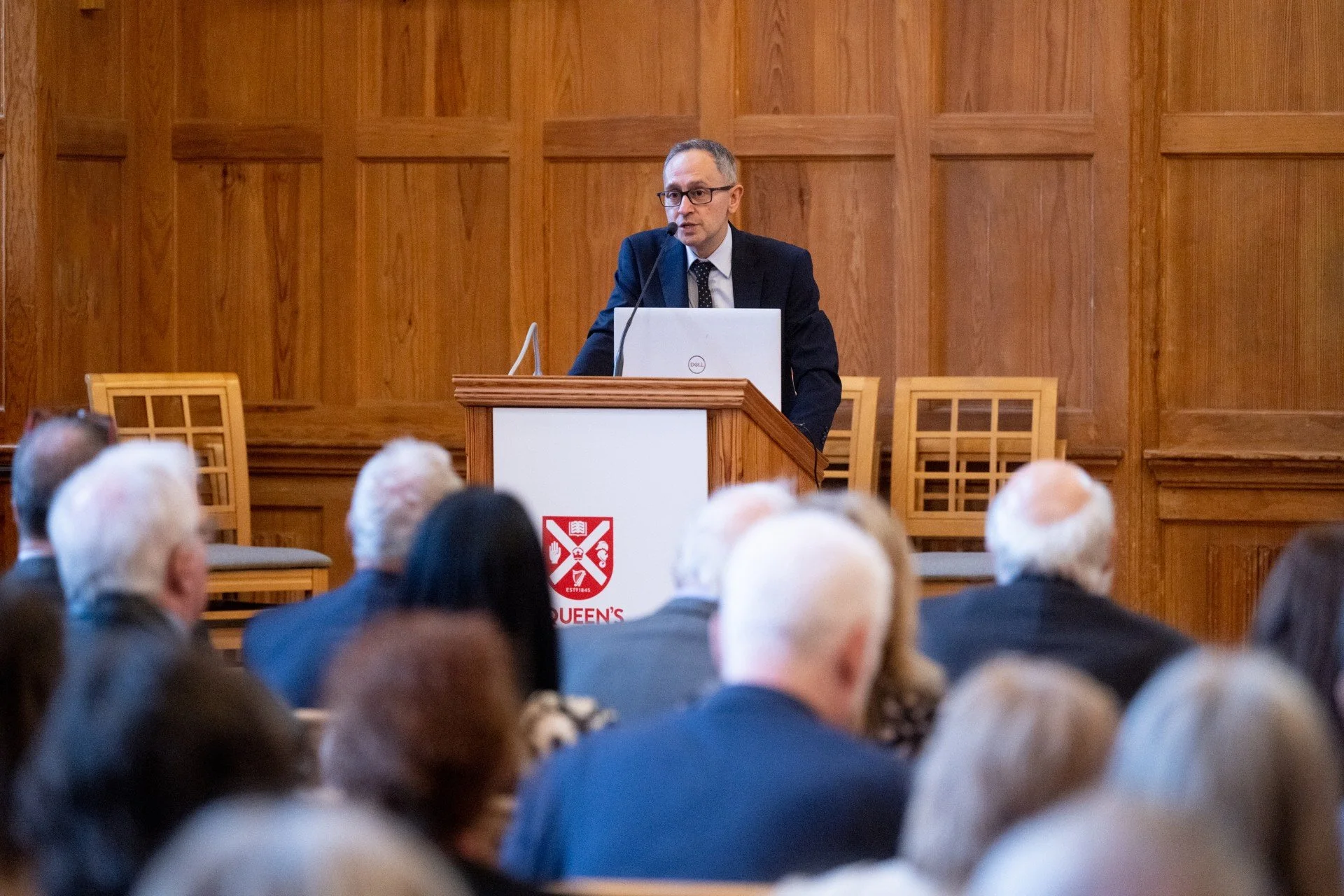 A man in a suit and glasses giving a lecture at a podium with a laptop, in front of an audience in a wood-paneled room with a university crest on the front.