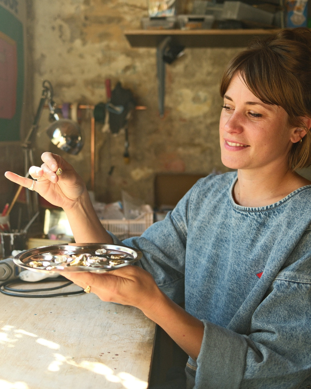 Une femme souriante tient une petite assiette avec des bijoux, examinant un bijou. Elle porte un sweat en jean, dans un atelier avec un mur en pierre et des outils en arrière-plan.