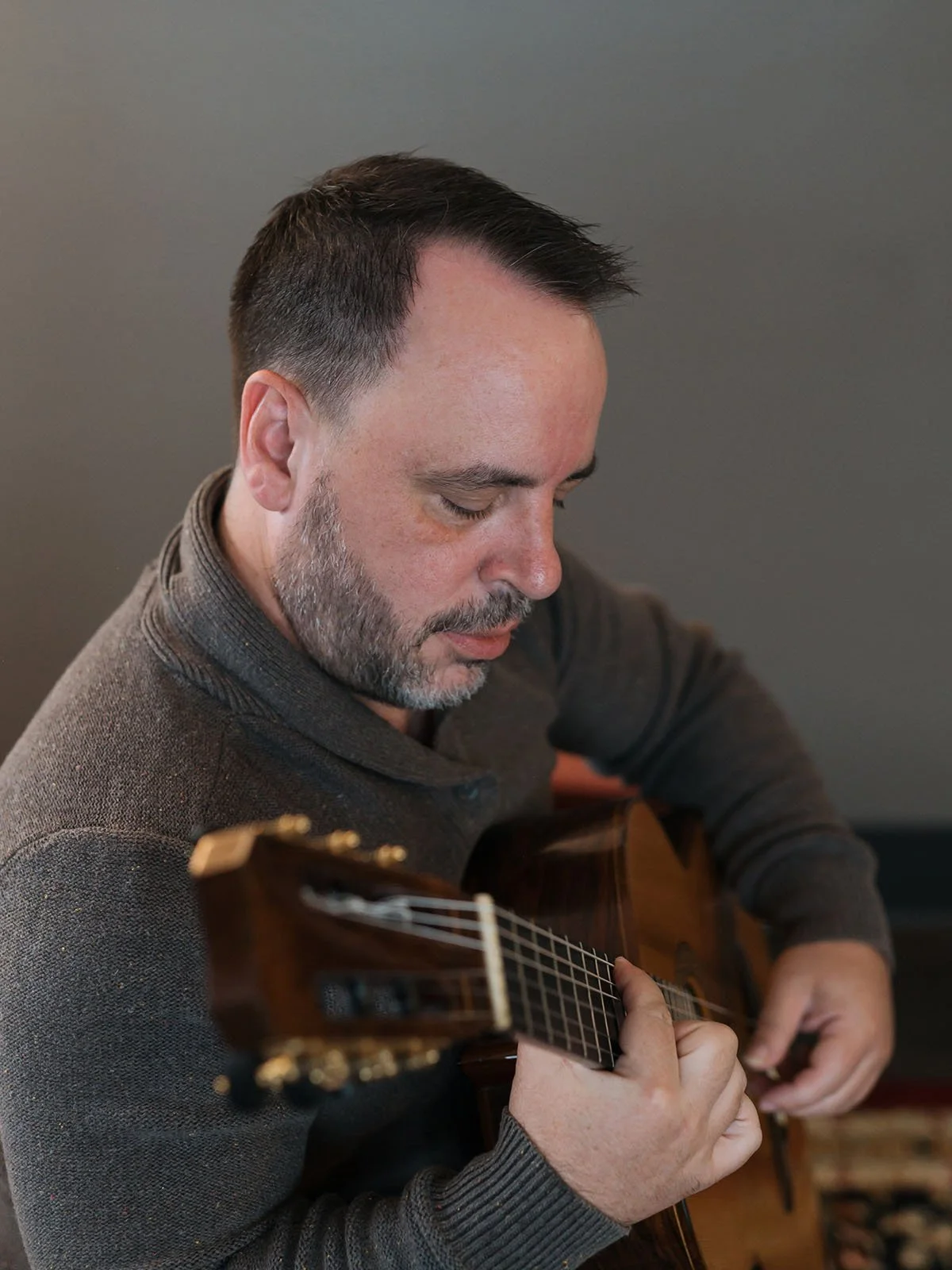 A man with dark hair and a gray beard playing an acoustic guitar with focused expression.