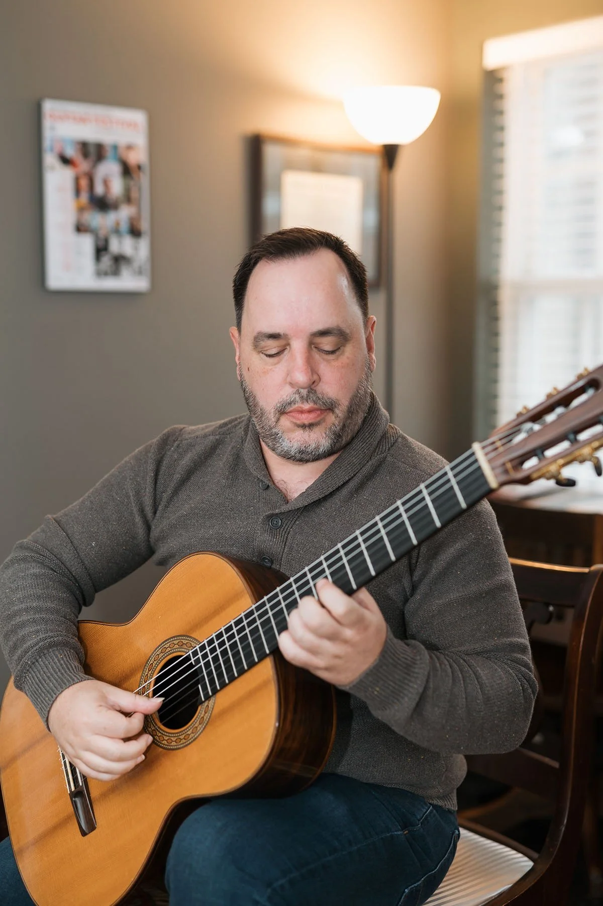 A man with a beard and closed eyes playing an acoustic guitar indoors near a window with blinds, a framed picture on the wall, and a standing lamp.