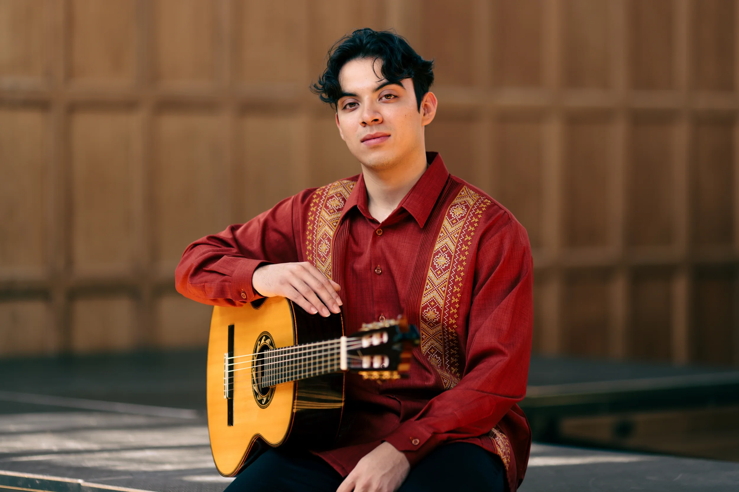 A young man with black hair in a middle part, wearing a red guayabera shirt and holding a classical guitar