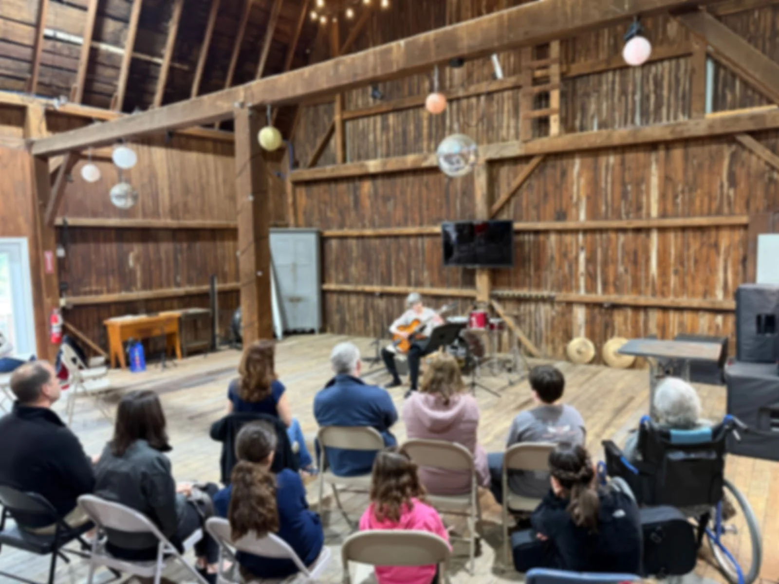 People watching a live music performance in a rustic, barn-like setting with wooden walls and ceiling. The performer is playing an acoustic guitar, and the audience is seated on folding chairs.