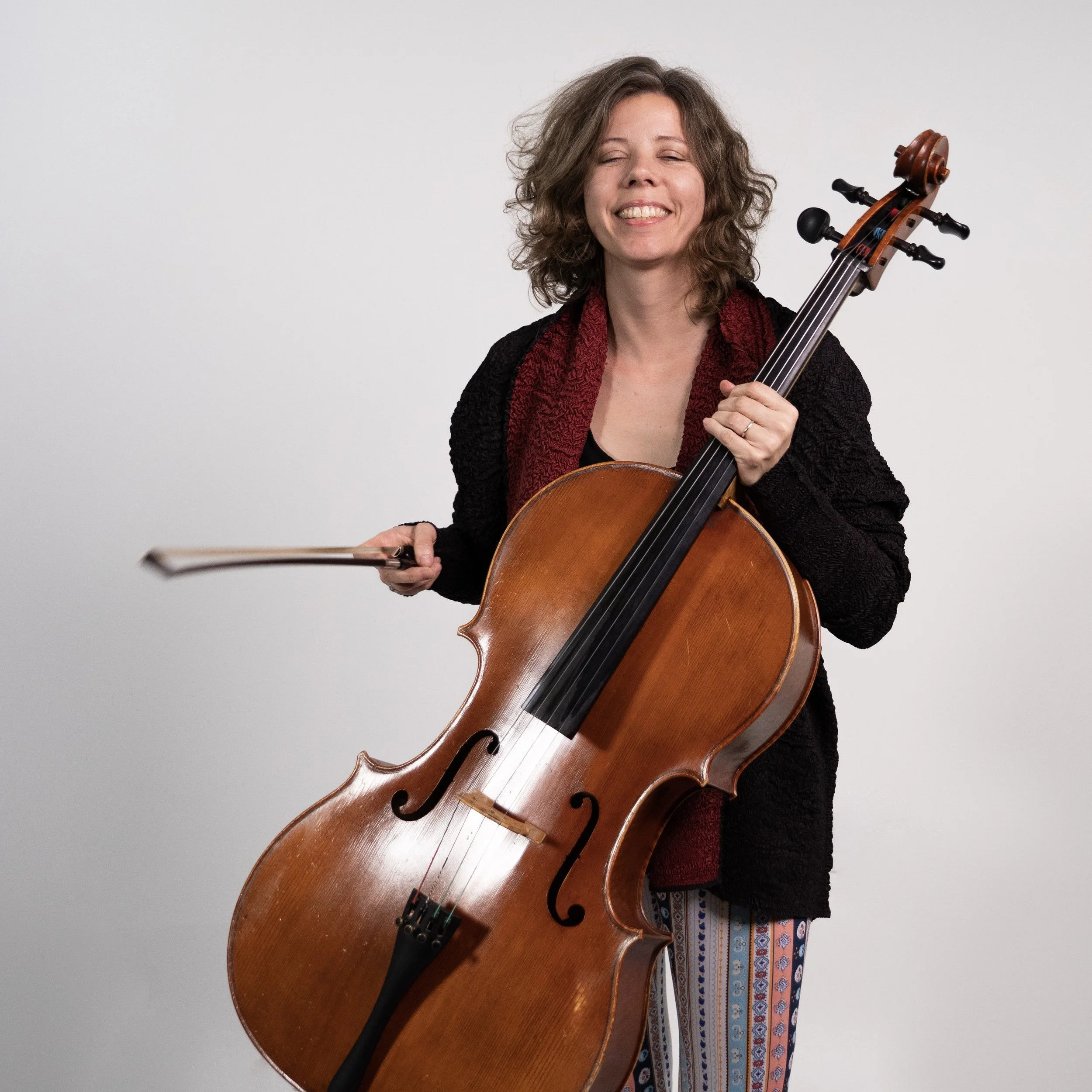A woman holding a cello, smiling with her eyes closed holding her bow out