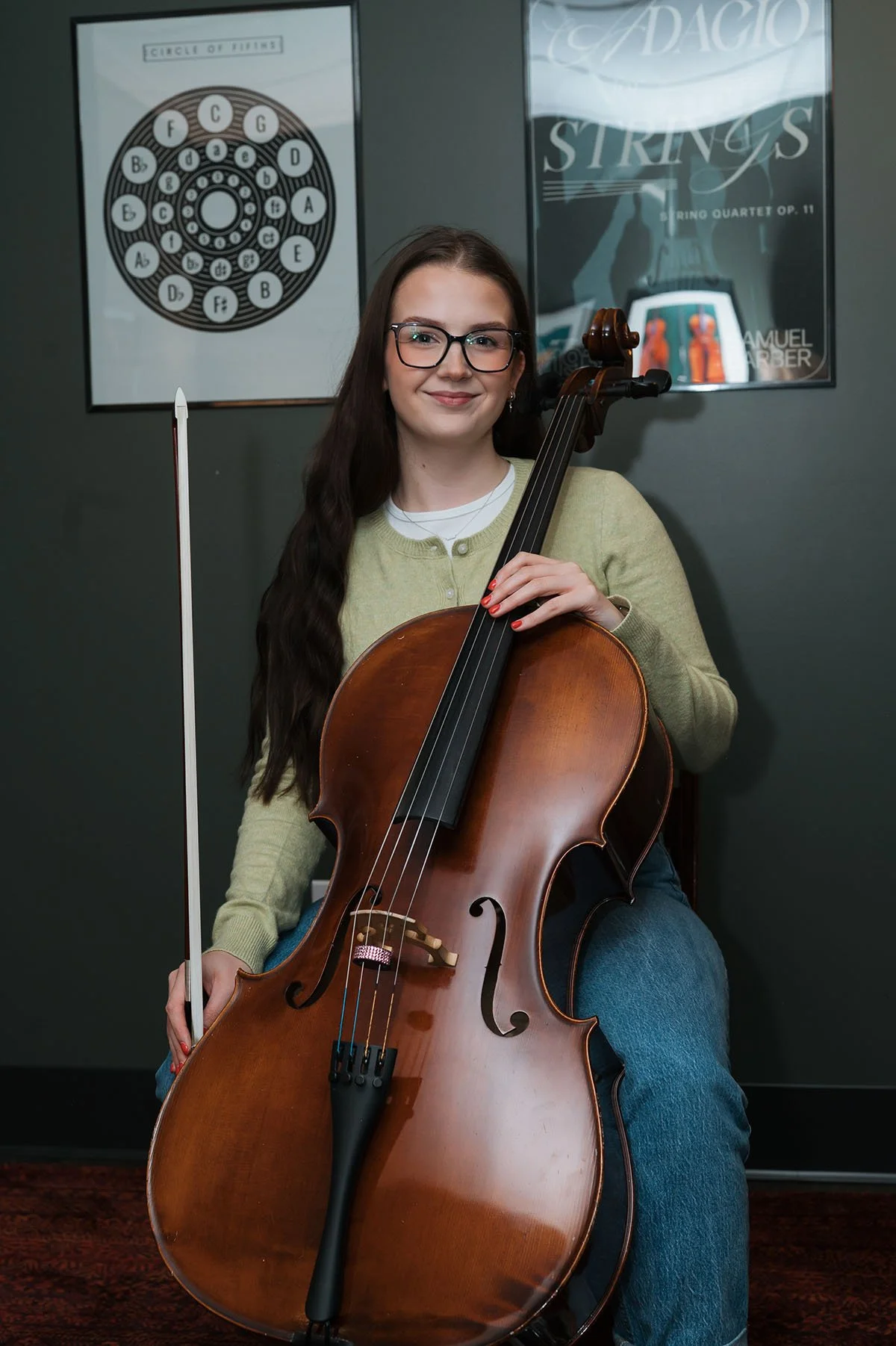Young woman with glasses sitting and holding a cello, in a music room with framed posters on the wall, including one with a circle of fifths and another with 'Adagio Strings'.