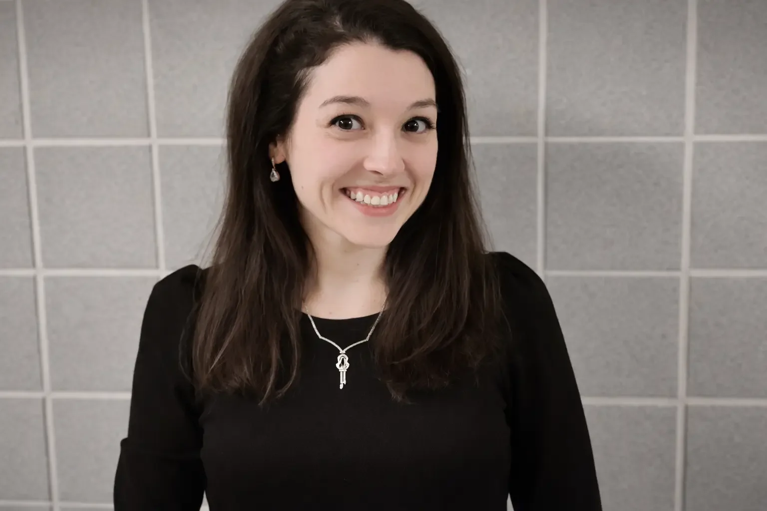 A young woman in all black with black hair and a gold necklace smiling.