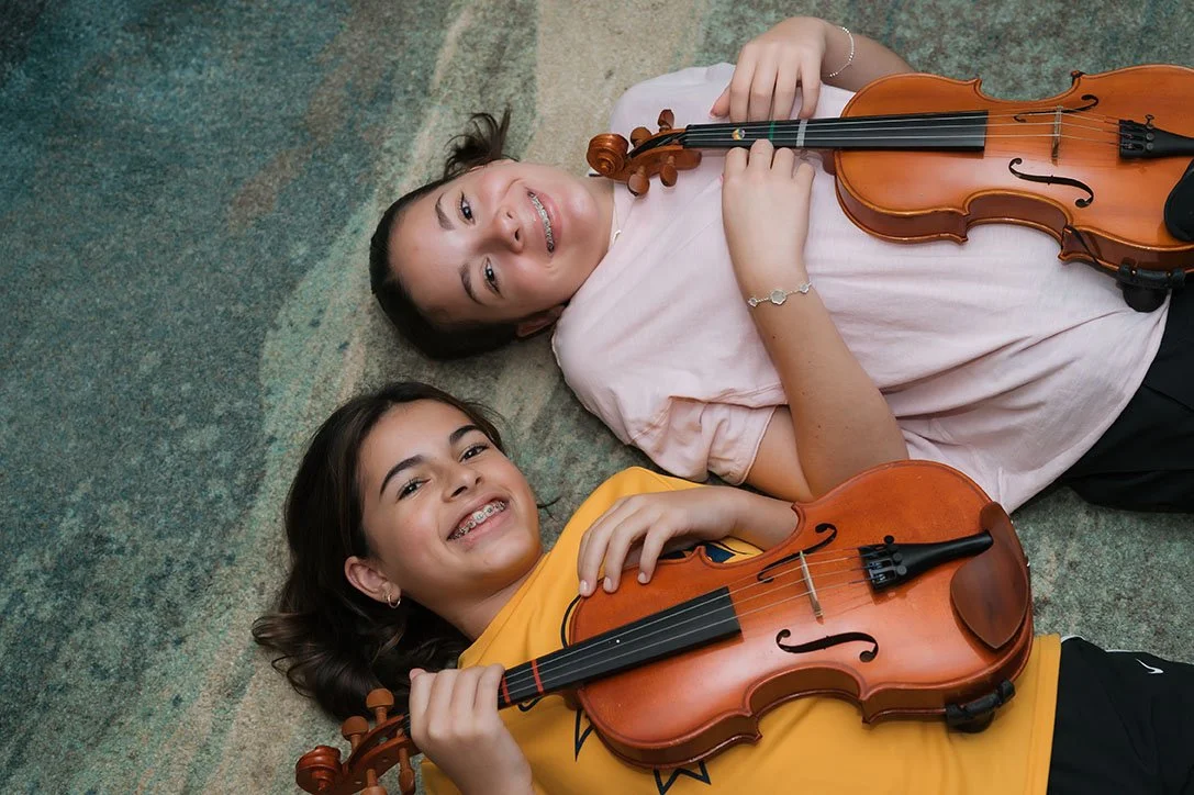 Two young girls lying on a carpeted floor, holding violins and smiling at the camera.