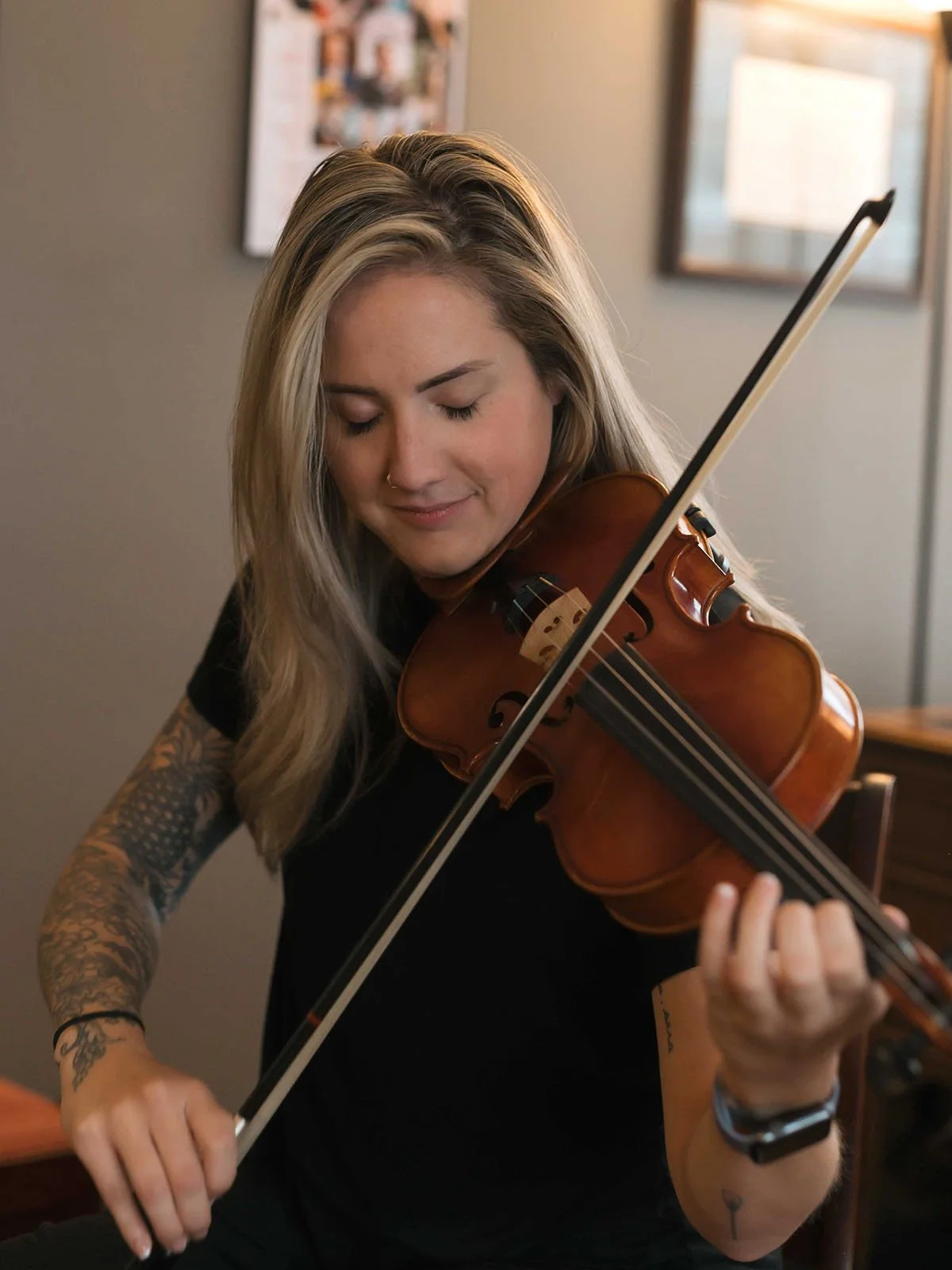 A young woman with blonde hair playing a violin, eyes closed, in a room with framed pictures on the wall.