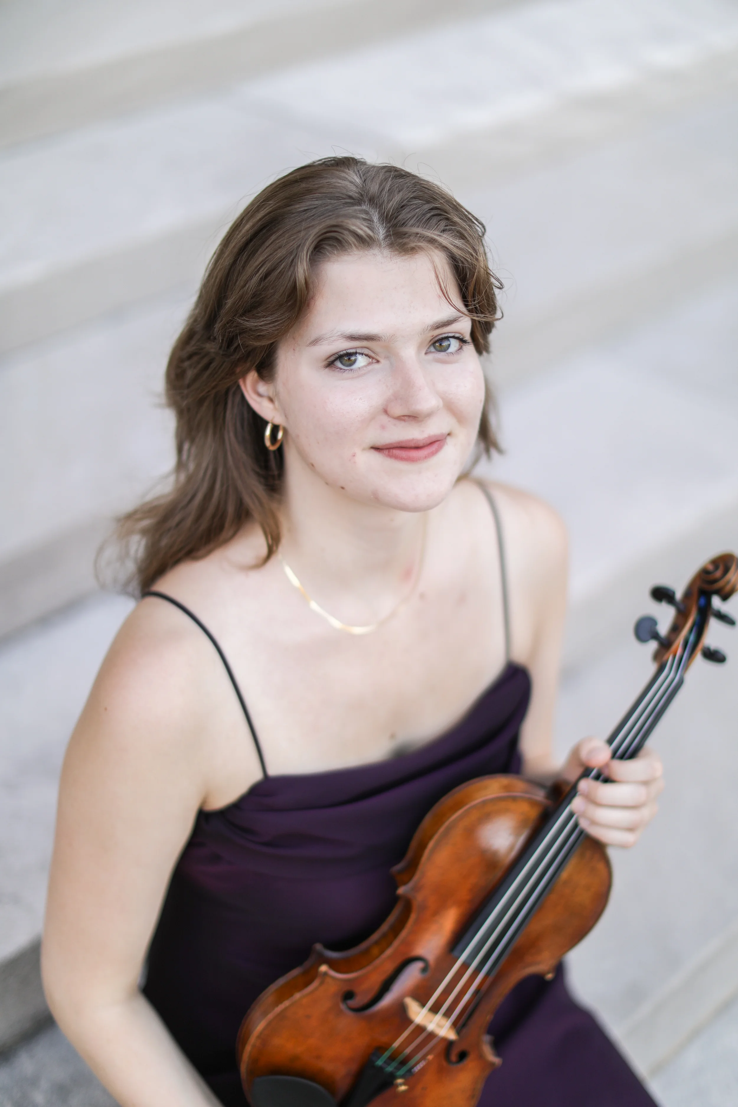 A young woman in a black dress with dirty blonde hair and blue eyes, smiling while holding a violin