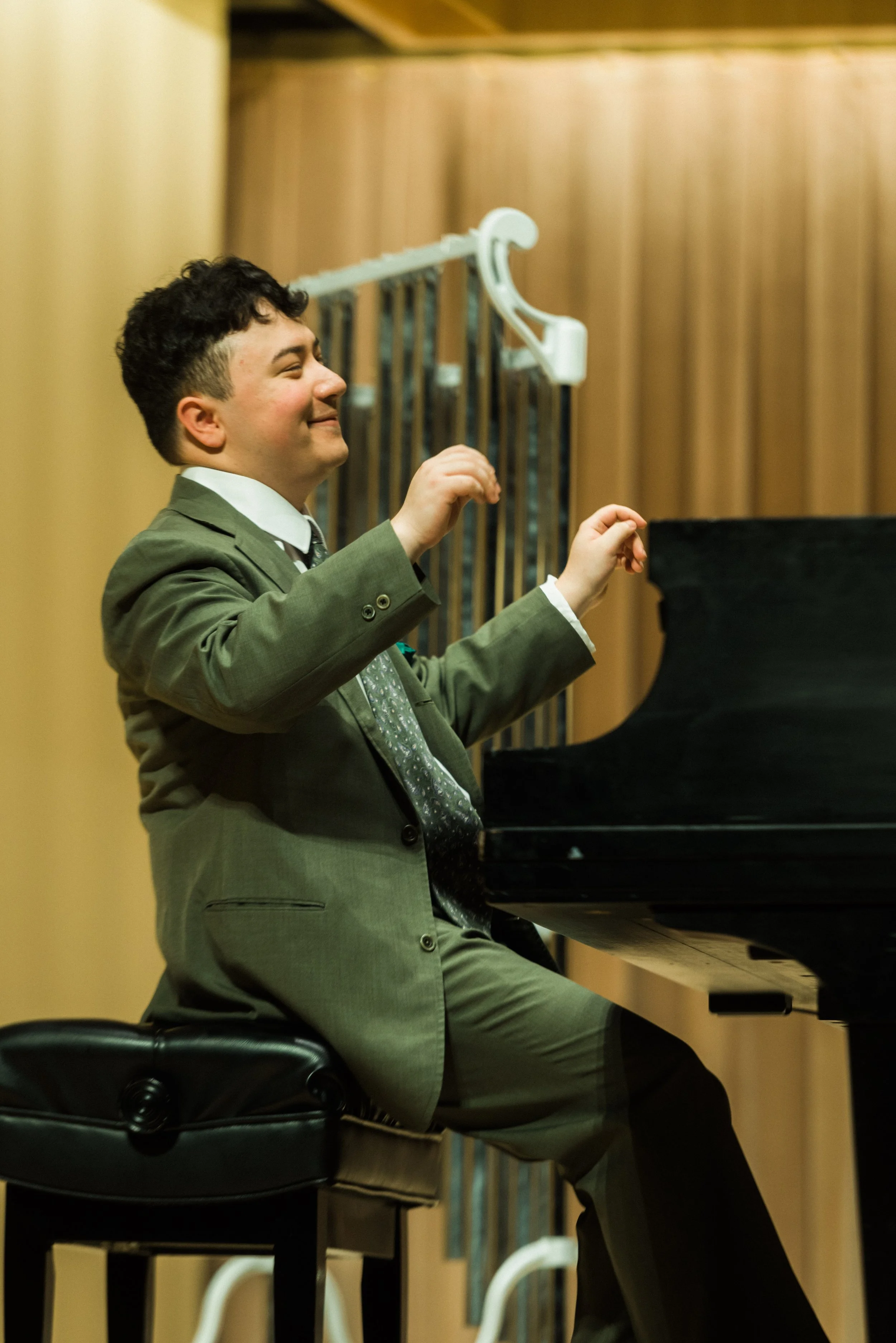 A young man in a green suit with his hands up in the air and smiling with his eyes closed while sitting at a piano.
