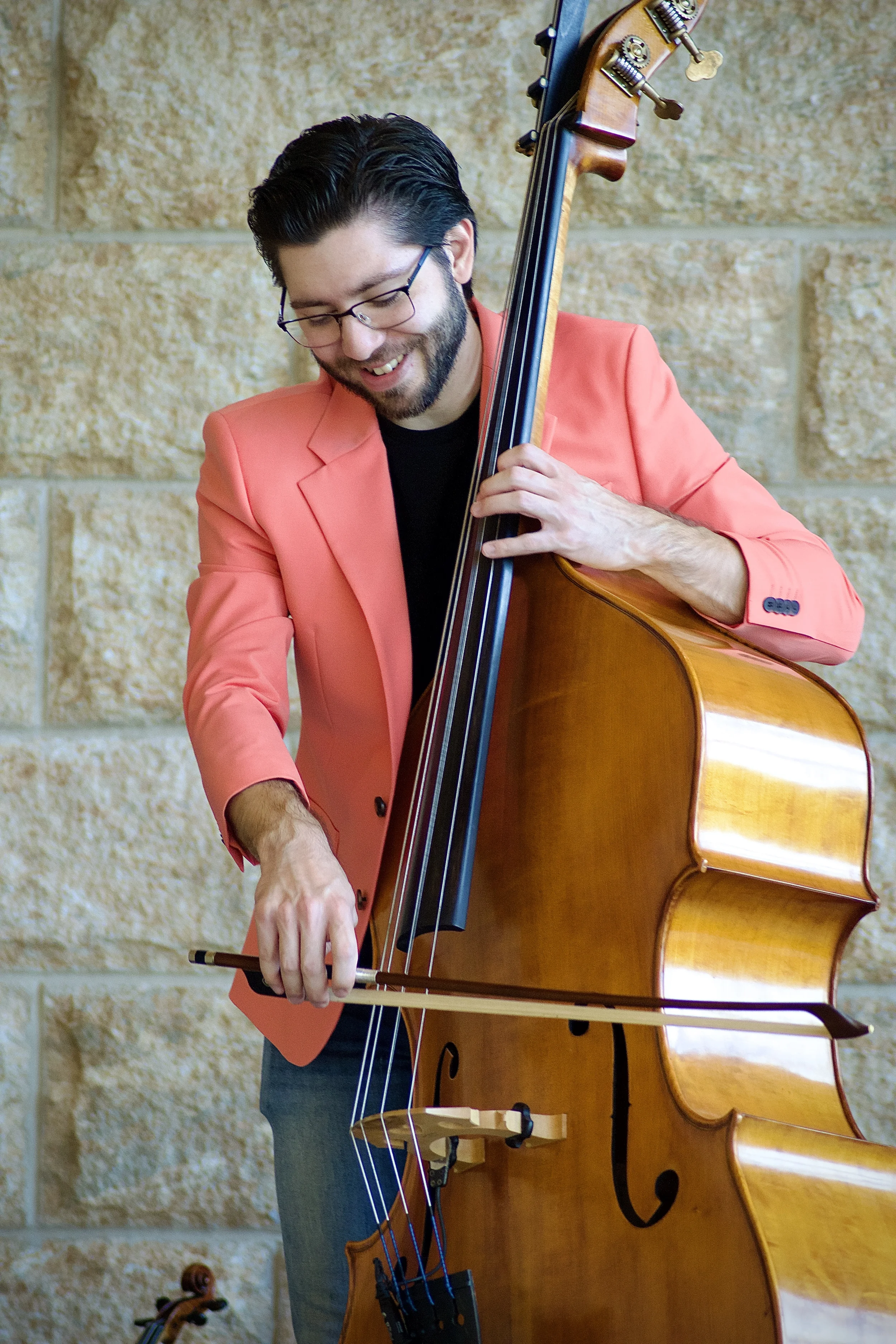 A young man with black hair and glasses, wearing a pink blazer and smiling while playing the double bass.