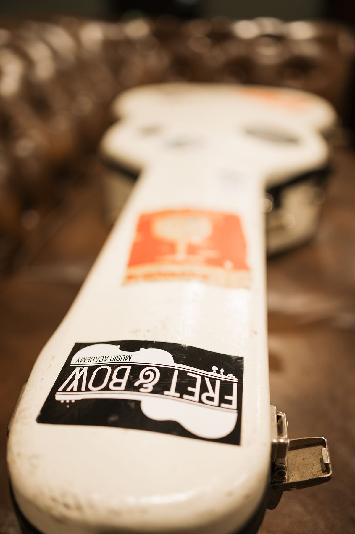 Close-up of a guitar case with stickers, including a black one that reads 'FREEL & BOW MUSIC ACADEMY,' and a blurred background of chairs.