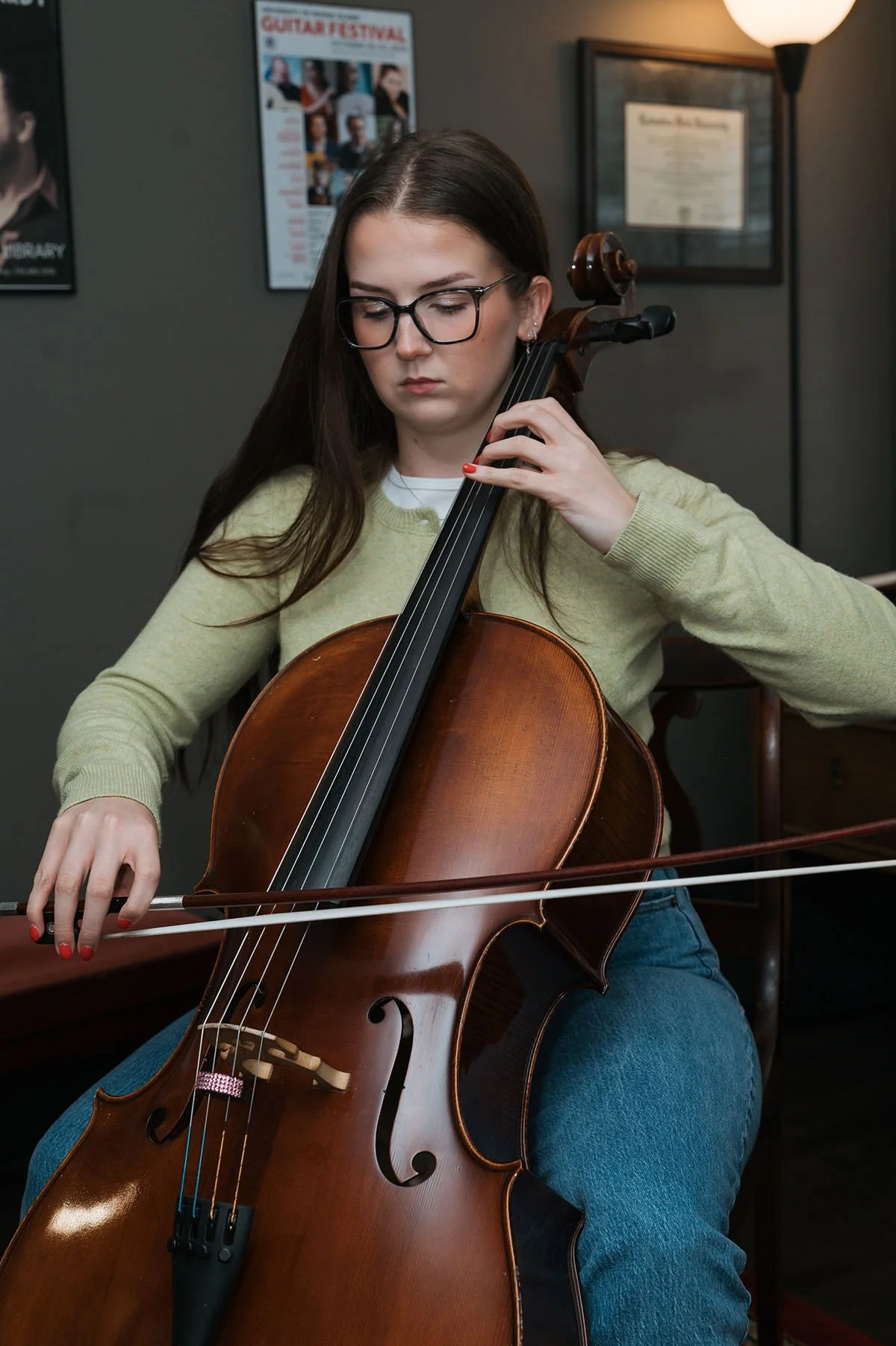 Young woman with glasses playing the cello indoors, sitting on a chair.