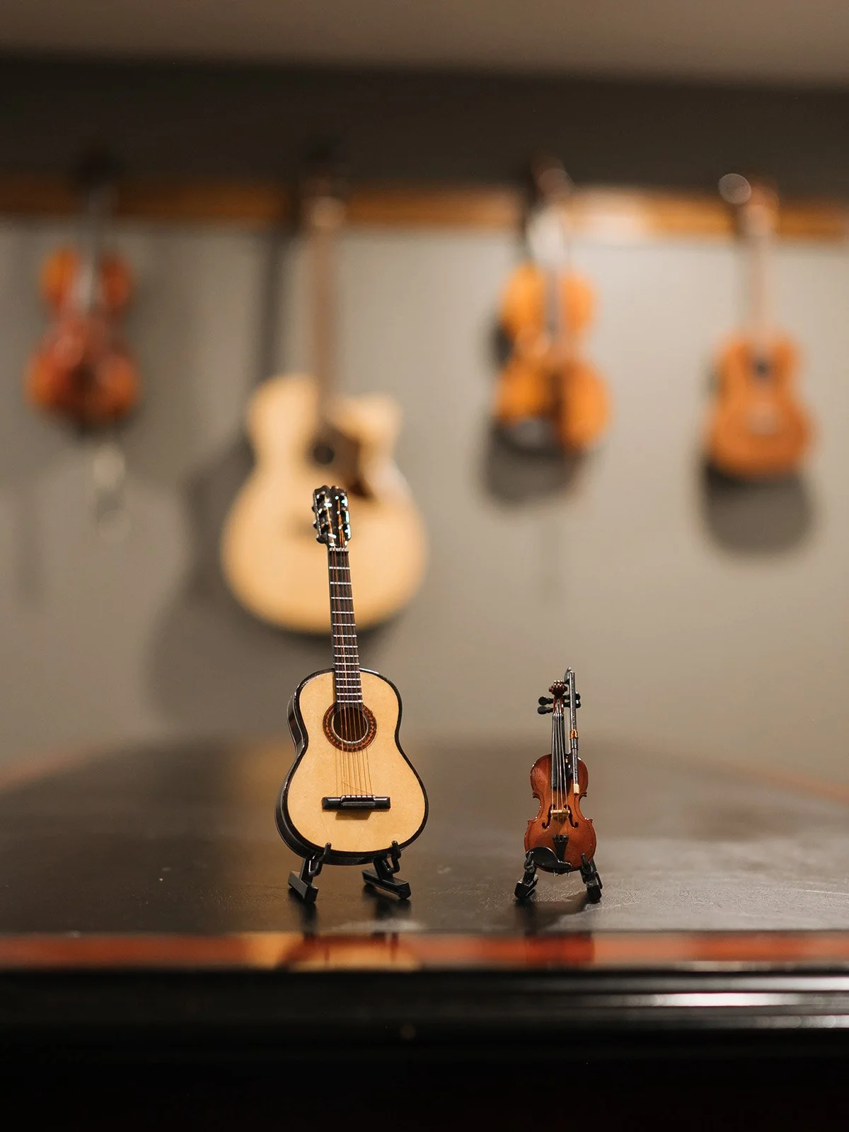 Two small toy string instruments, a guitar and a violin, placed on a table in front of a blurred background of hanging guitars.