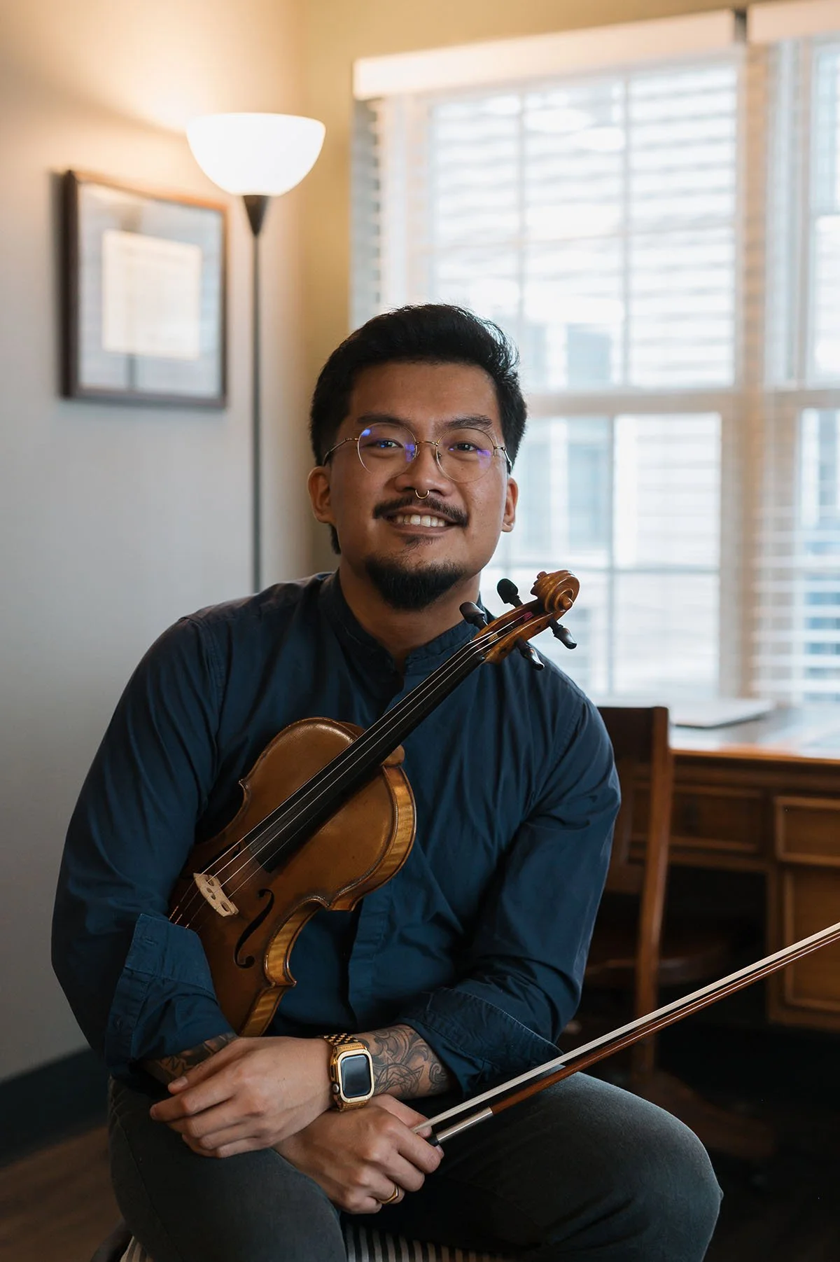 A man with glasses, a nose ring, and a tattoo on his arm, holding a violin and a bow, sitting in a room with a window with blinds, a desk, and a framed certificate, smiling at the camera.