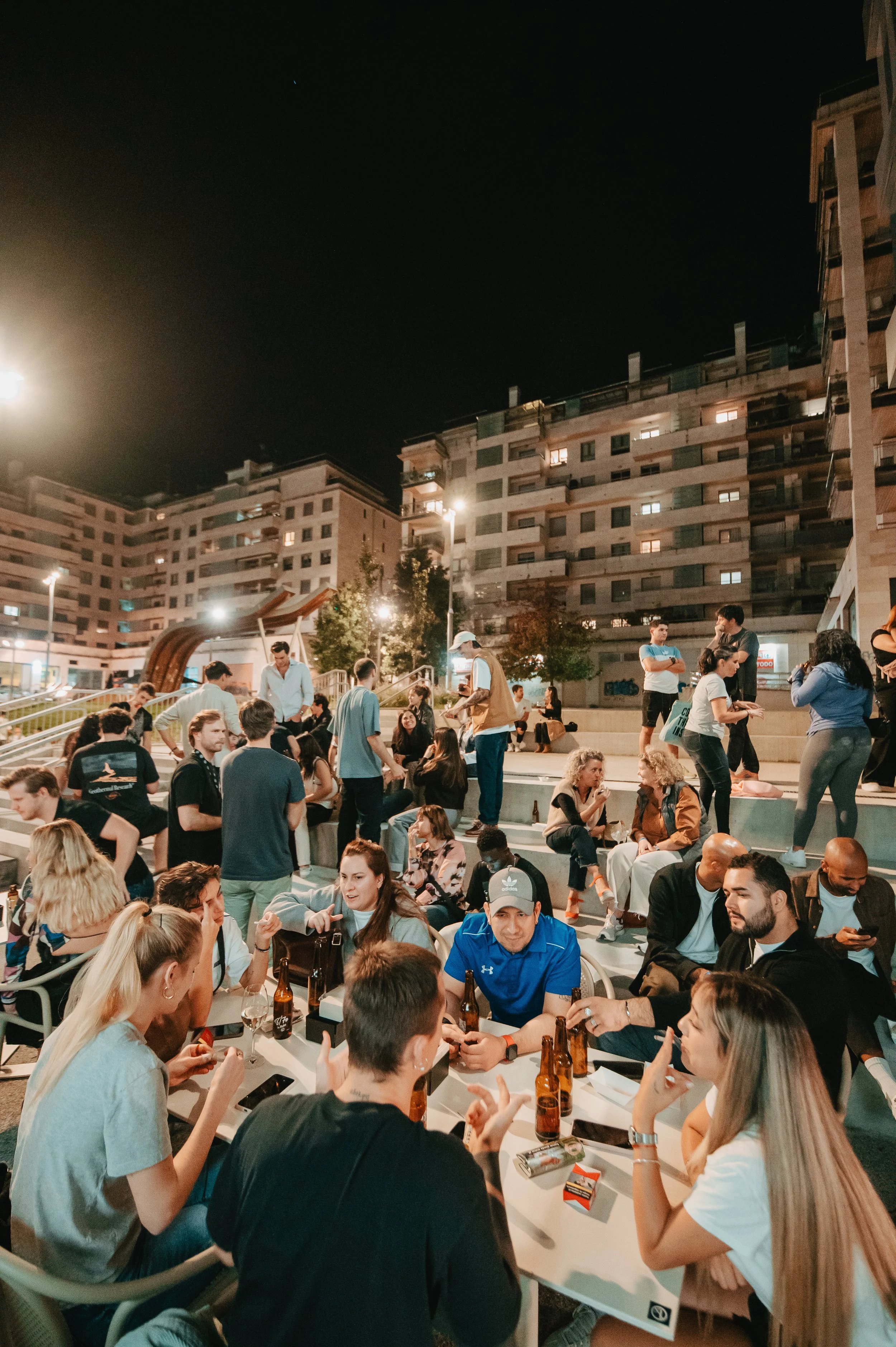 Grupo de personas conversando y socializando en un espacio público al aire libre por la noche, con edificios y luces en el fondo.