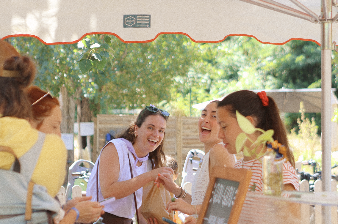 Grupo de mujeres sonriendo y charlando en un ambiente al aire libre bajo una sombrilla, en un día soleado.