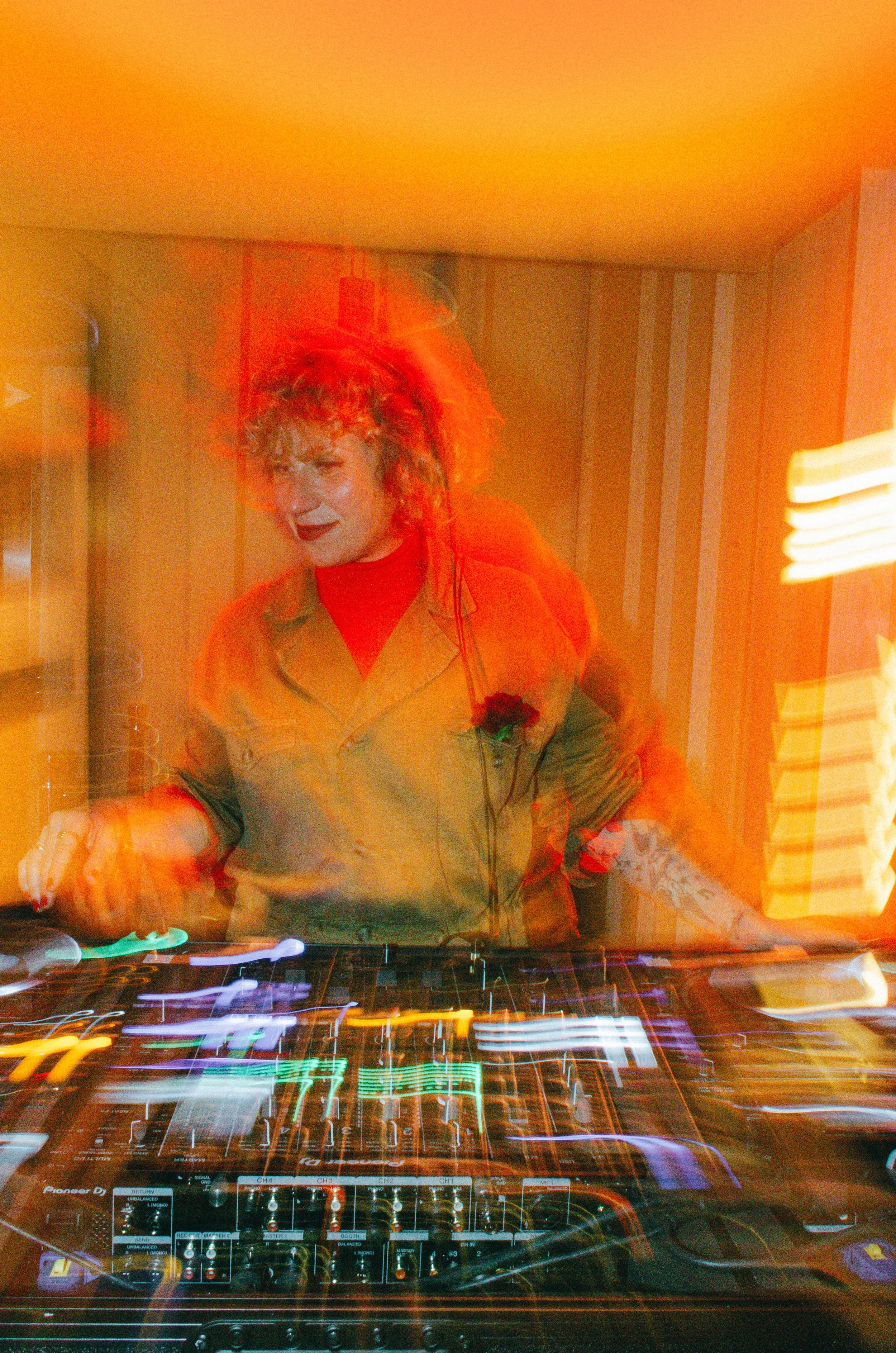 Un DJ tocando en una cabina con luces y equipo de mezclas, con un fondo de paredes con rayas.