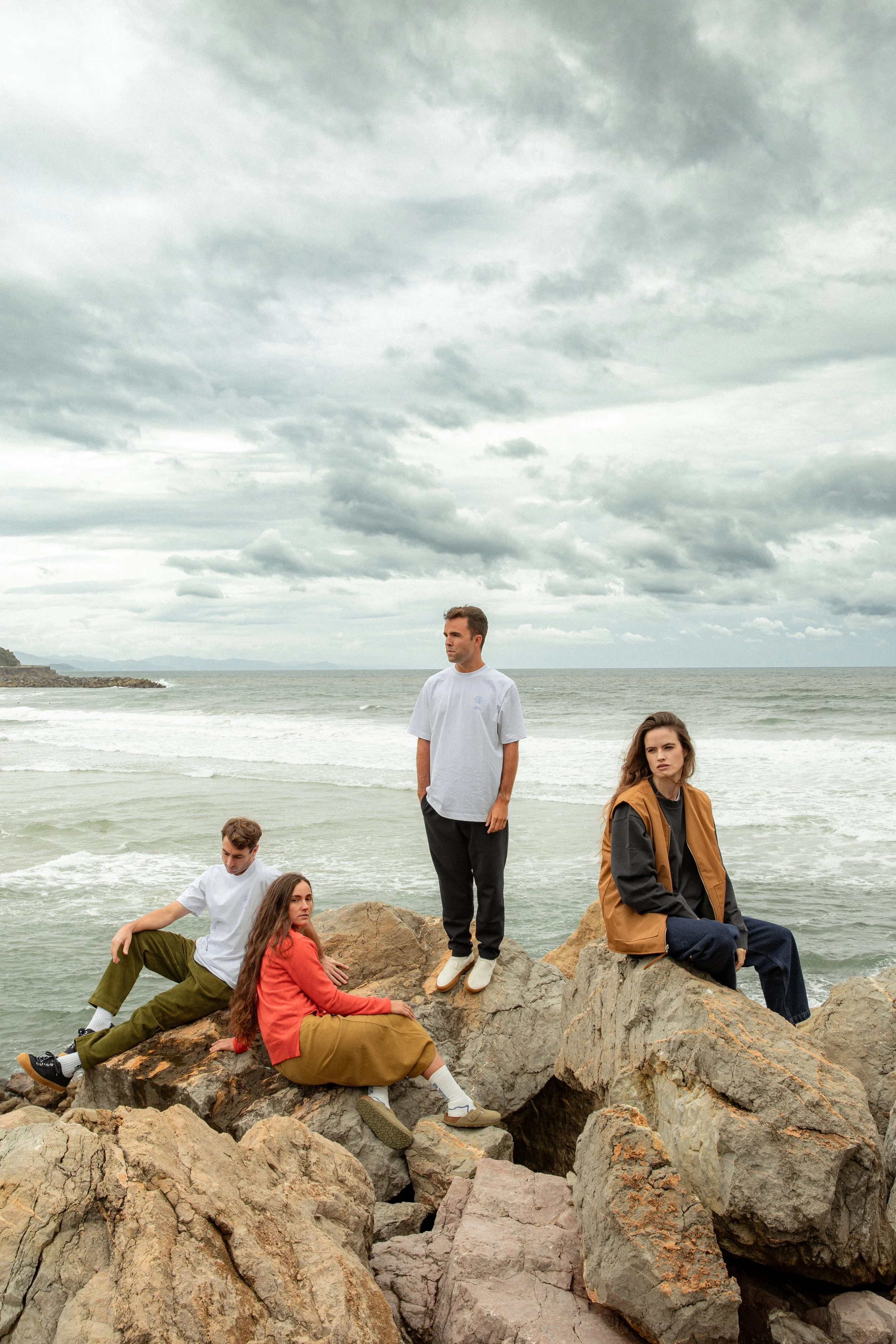 Un grupo de cinco personas en una playa con rocas y el océano, durante un día nublado. Algunas personas están sentadas en las rocas, mientras otras están de pie, observando o posando para la foto.