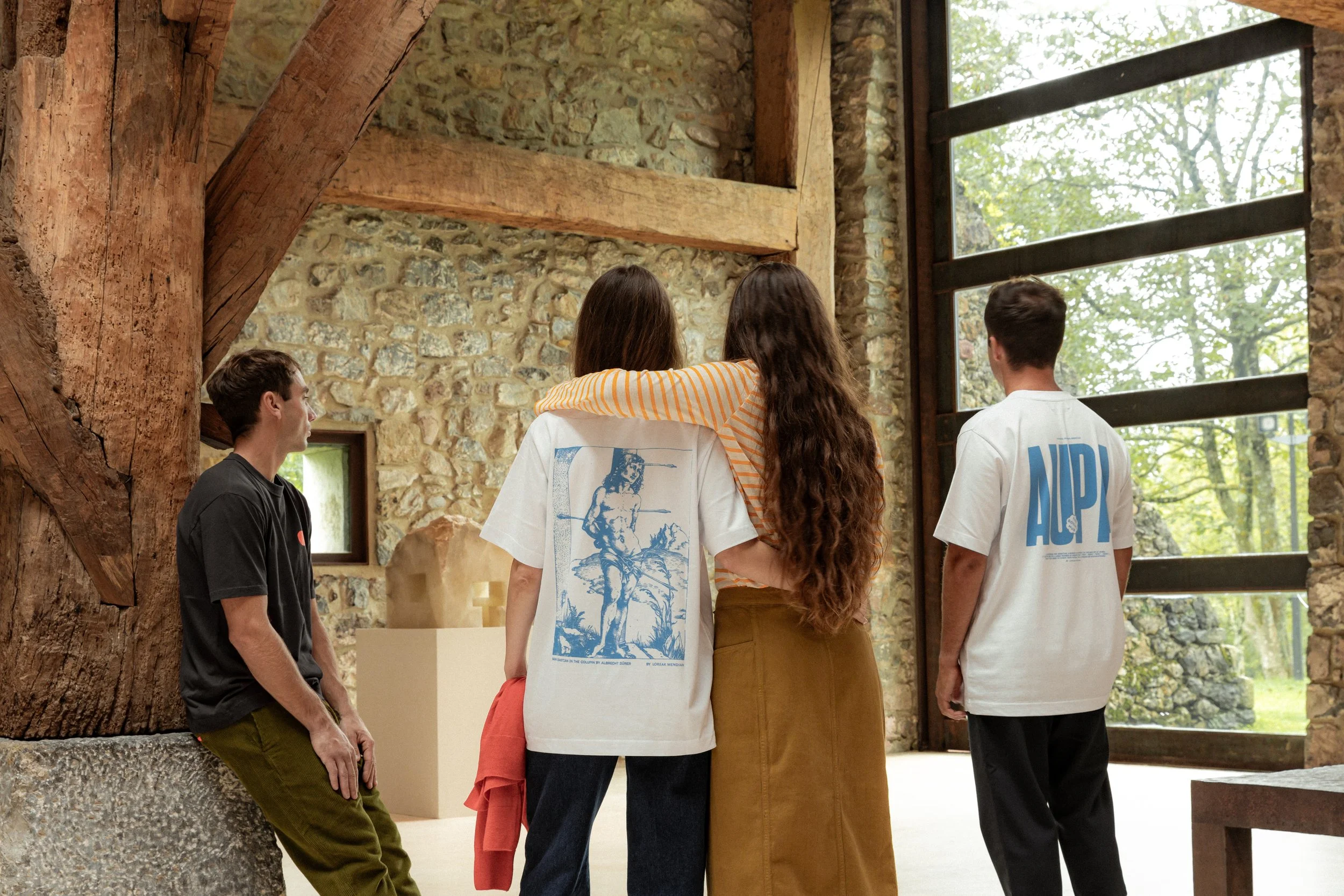 Grupo de jóvenes de espaldas, en una sala con paredes de piedra y ventanales grandes con vista a árboles, observando una exhibición en una vitrina.