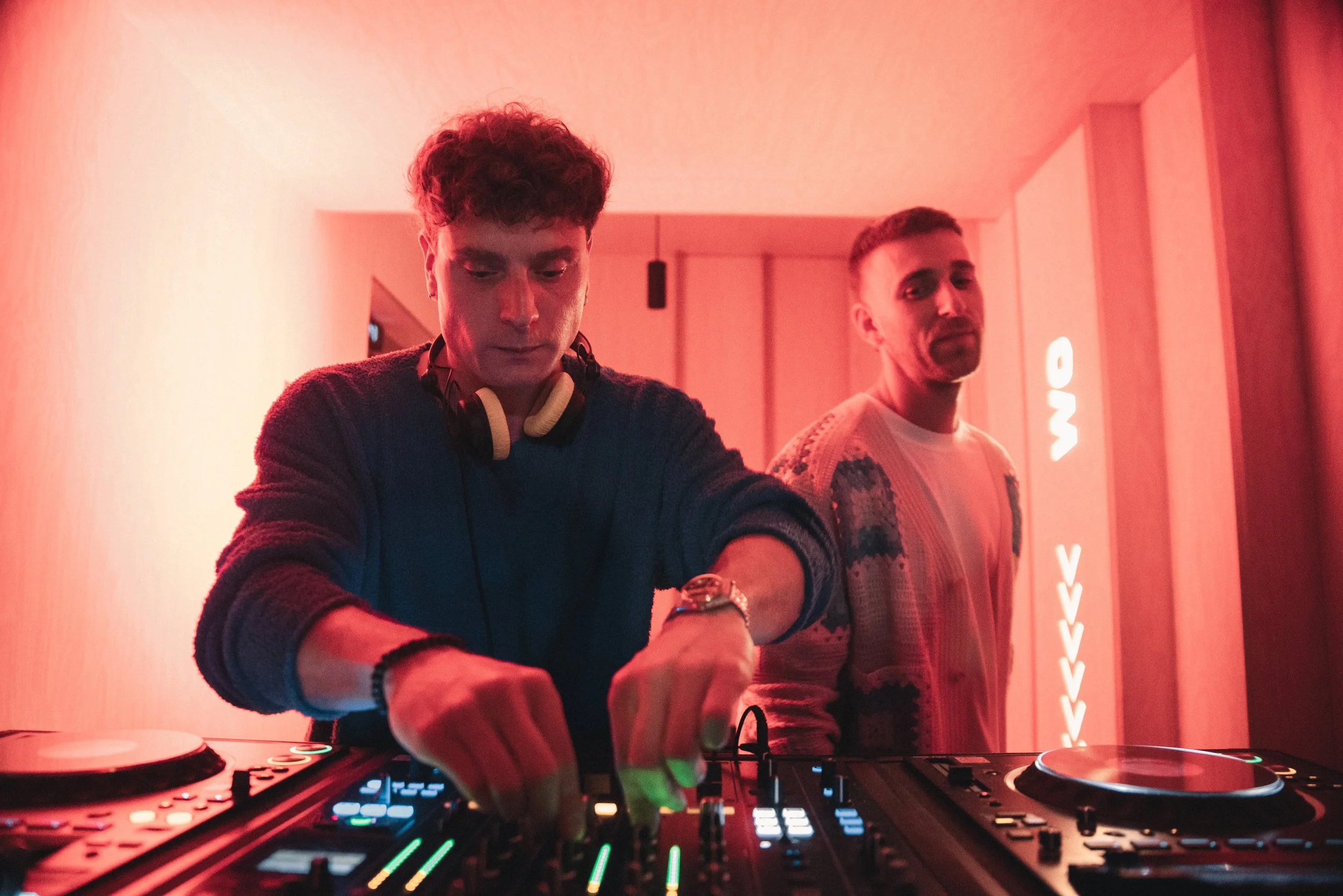 Dos hombres en un club nocturno con iluminación roja, uno es DJ usando audífonos y controlando la música, el otro observa.