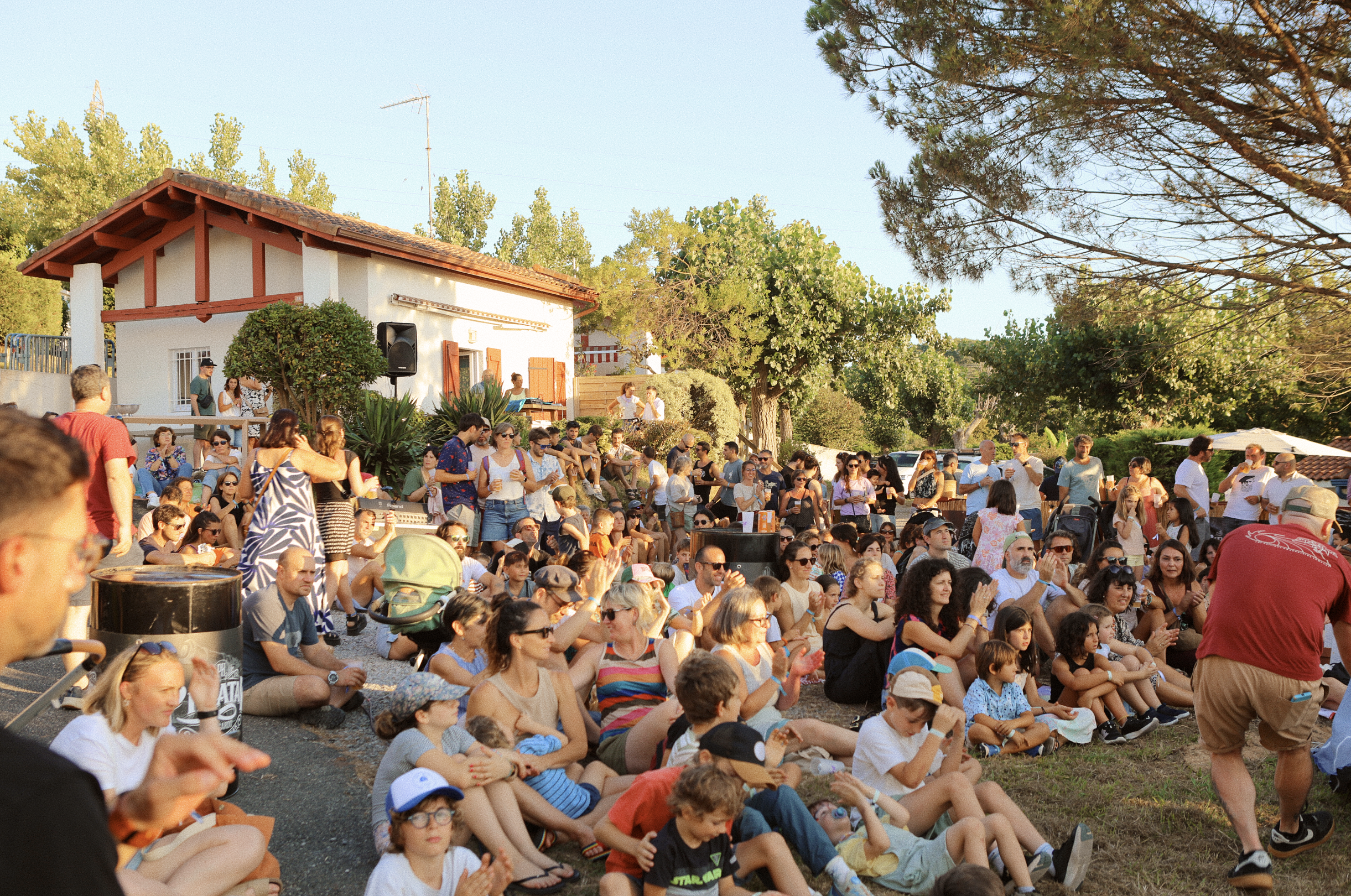 Grupo de personas sentado y de pie en un evento al aire libre en un día soleado, con árboles y una casa en el fondo.