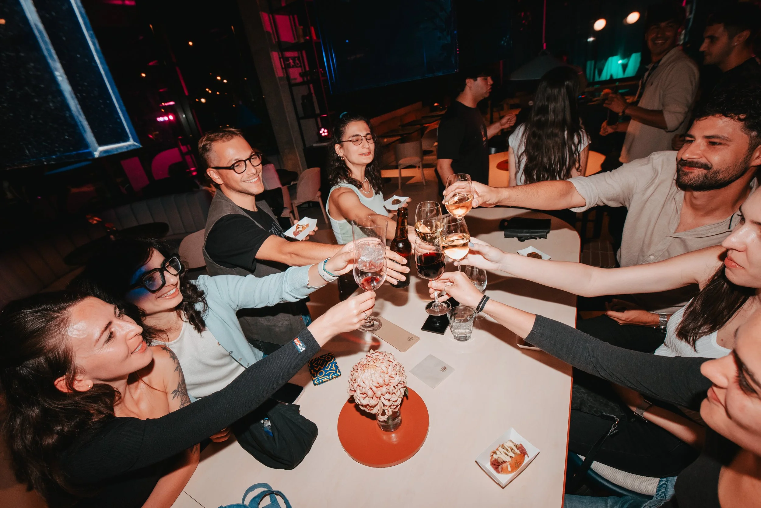 Grupo de personas en una fiesta en un restaurante, brindando con copas de vino y cerveza, algunos con ojos cerrados y sonriendo, en un ambiente oscuro con luces de neón.