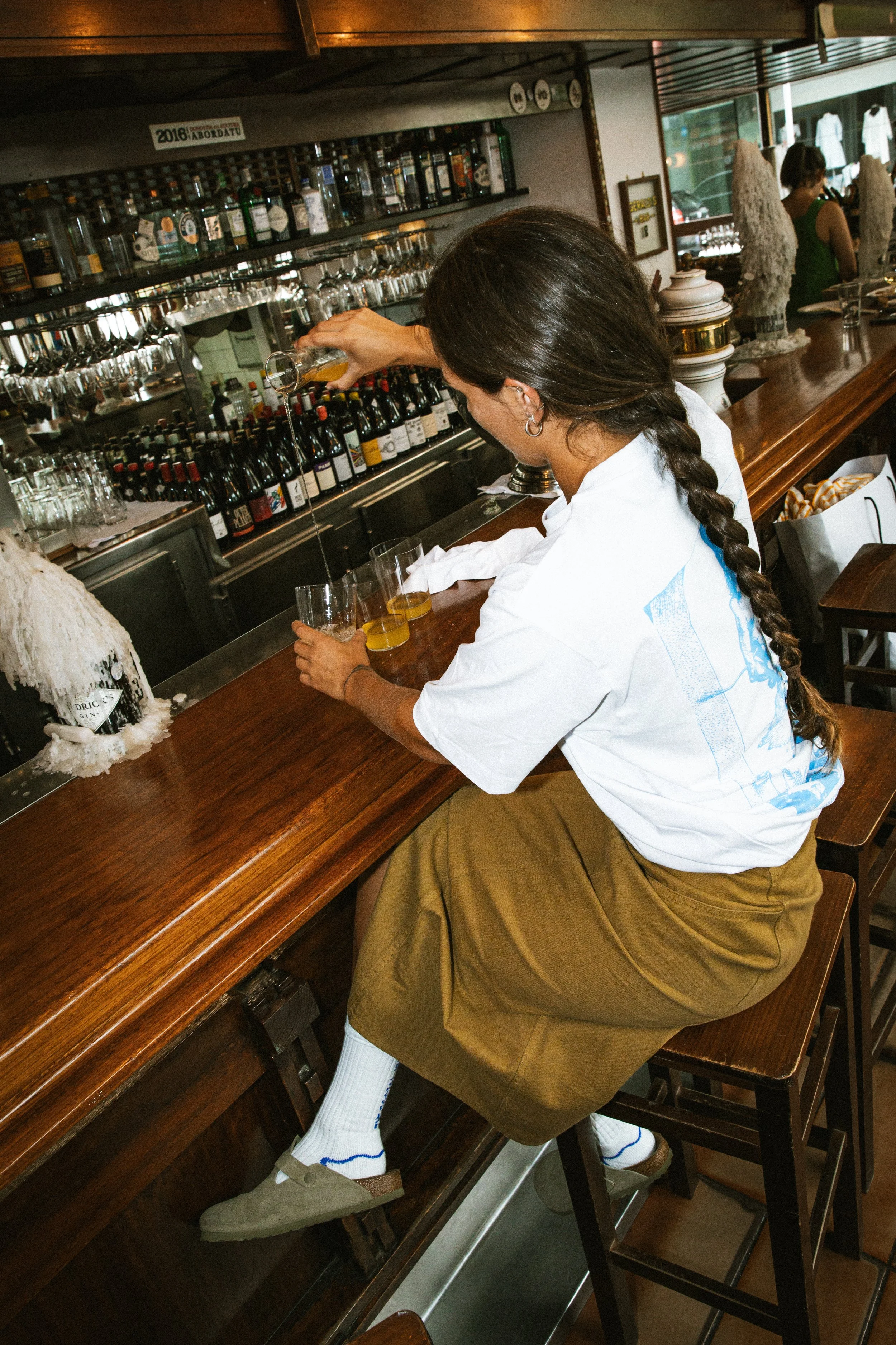 Persona sirviendo bebidas en un bar, vista lateral, con estanterías llenas de botellas y vasos, ambiente de cafetería o bar, decoraciones en el fondo, y otra persona en la esquina.