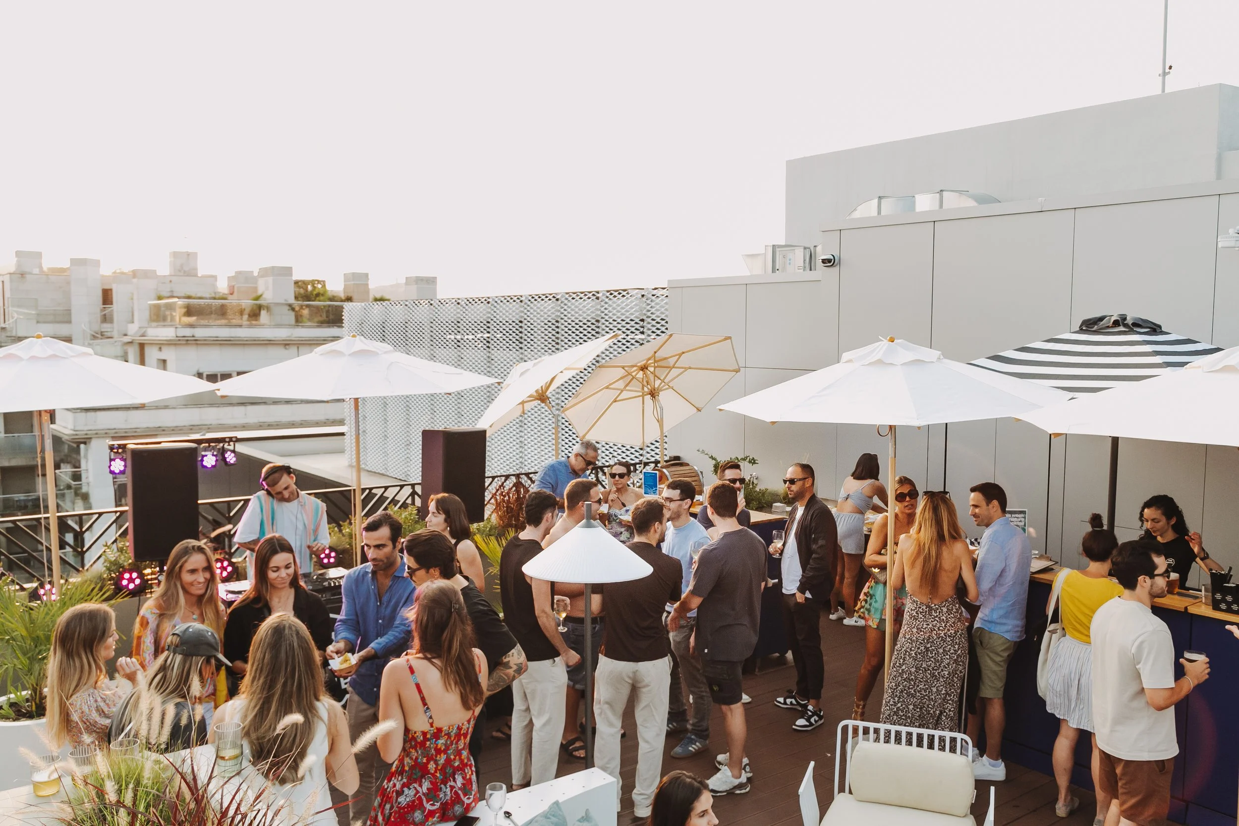 Superficie de terraza en la azotea con muchas personas disfrutando un evento social, con sombrillas blancas y decoraciones. En el fondo, edificios urbanos y cielo despejado.