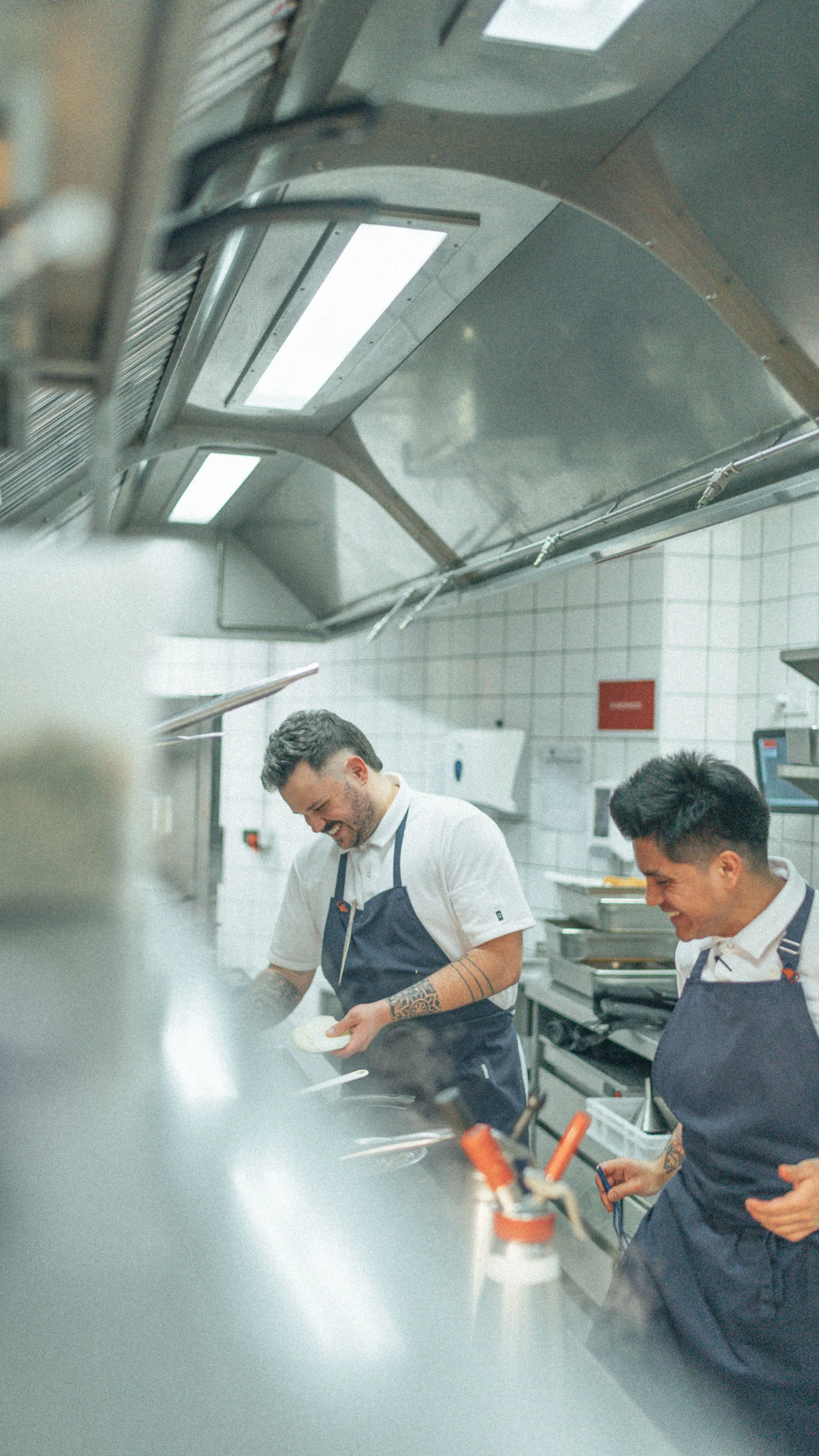 Dos hombres sonrientes trabajando en una cocina profesional, usando delantales oscuros, con equipo de cocina y una campana extractora sobre ellos.