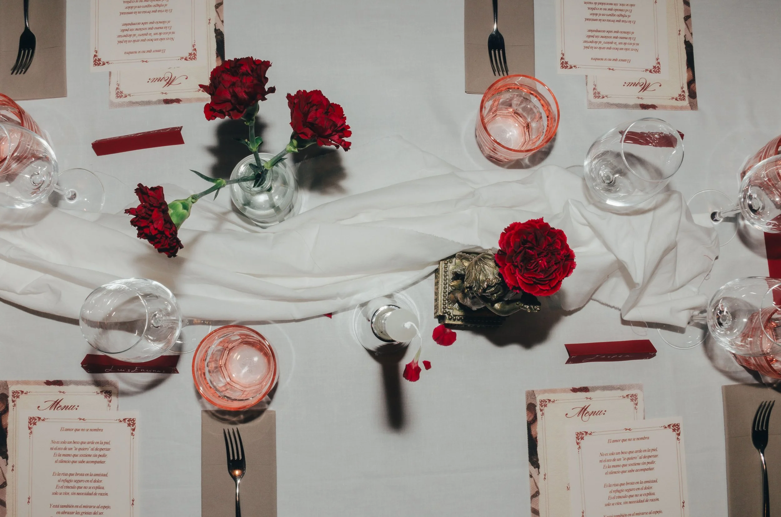Mesa decorada con flores rojas en pequeños jarrones, varias copas de vino o agua, menús en papel y cubiertos, sobre una mantelería blanca con detalles rojos.
